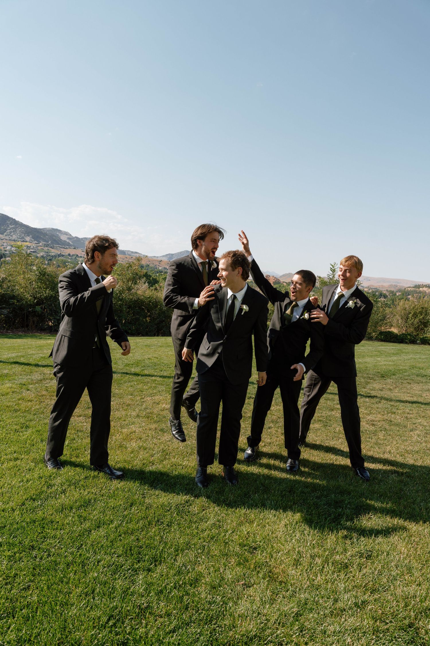 Groomsmen in dark suits celebrate and laugh together on lawn with mountains behind them.