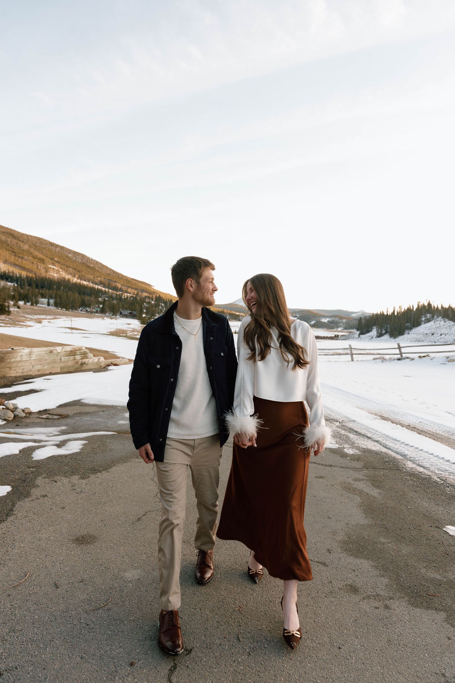 Couple walking hand in hand on a mountain path with snow and hills in background.