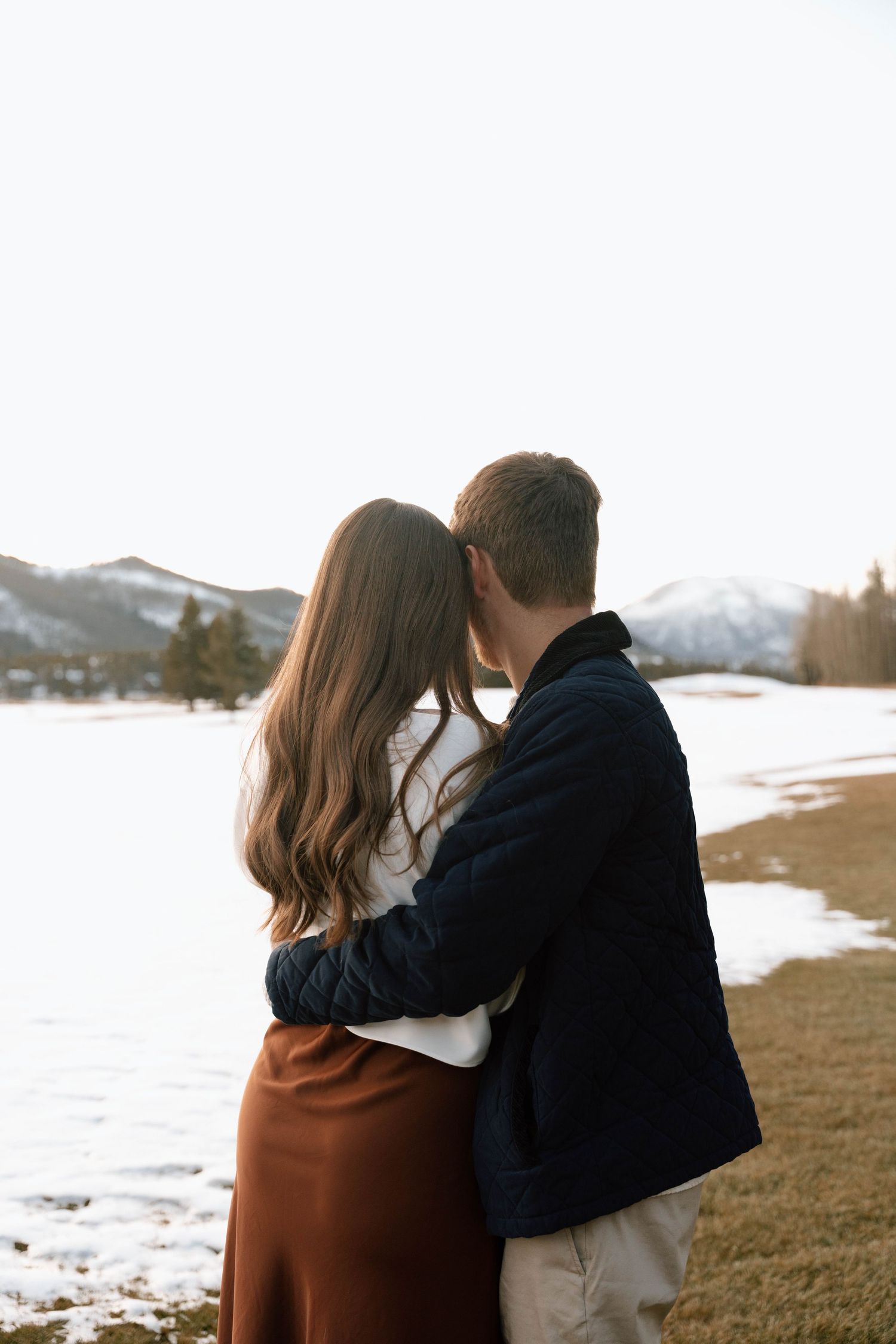 A couple stands together overlooking a snowy mountain vista at sunset.