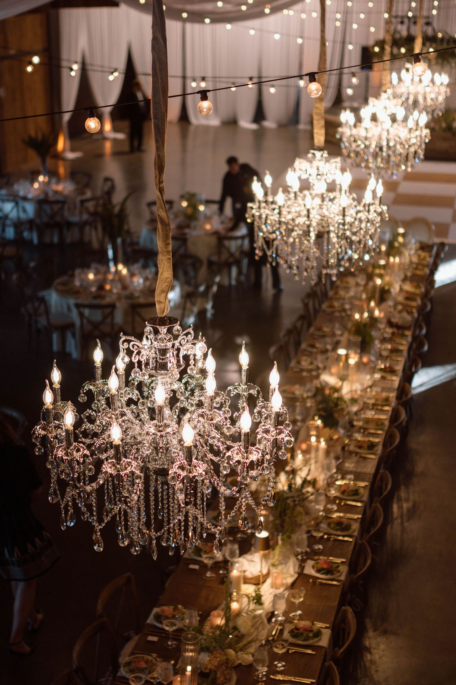 Elegant wedding reception with crystal chandeliers illuminating a long dining table set with candles and floral centerpieces.