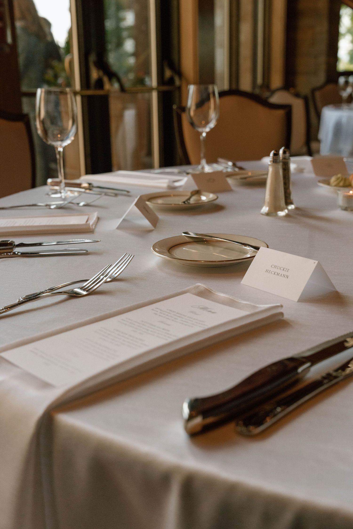Elegant restaurant table setting with white linen, silverware, wine glasses, menu cards, and place cards.