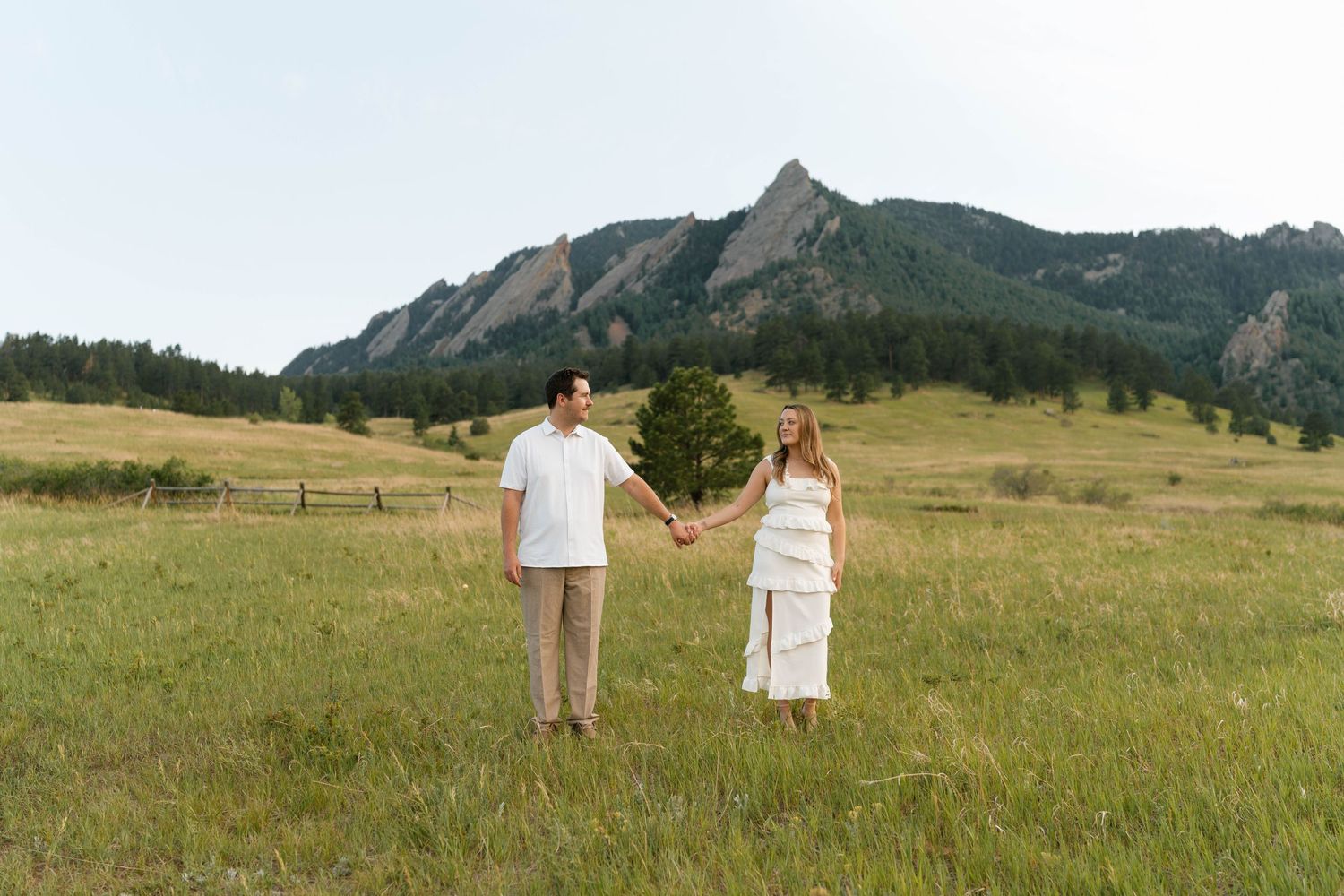Couple holds hands in green meadow with Colorado Flatirons mountain backdrop on sunny day.