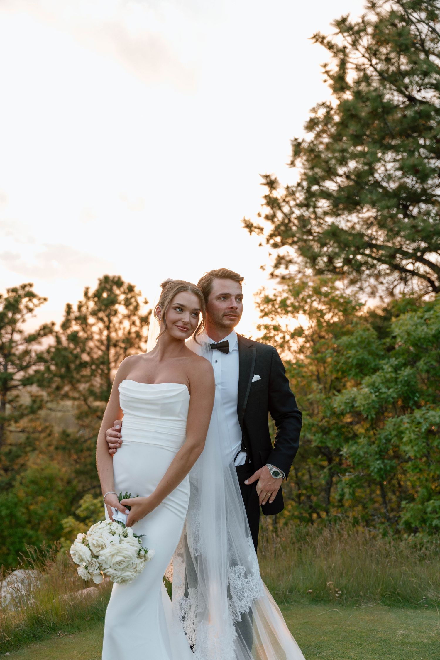 Romantic sunset wedding portrait with couple embracing on hilltop surrounded by trees.