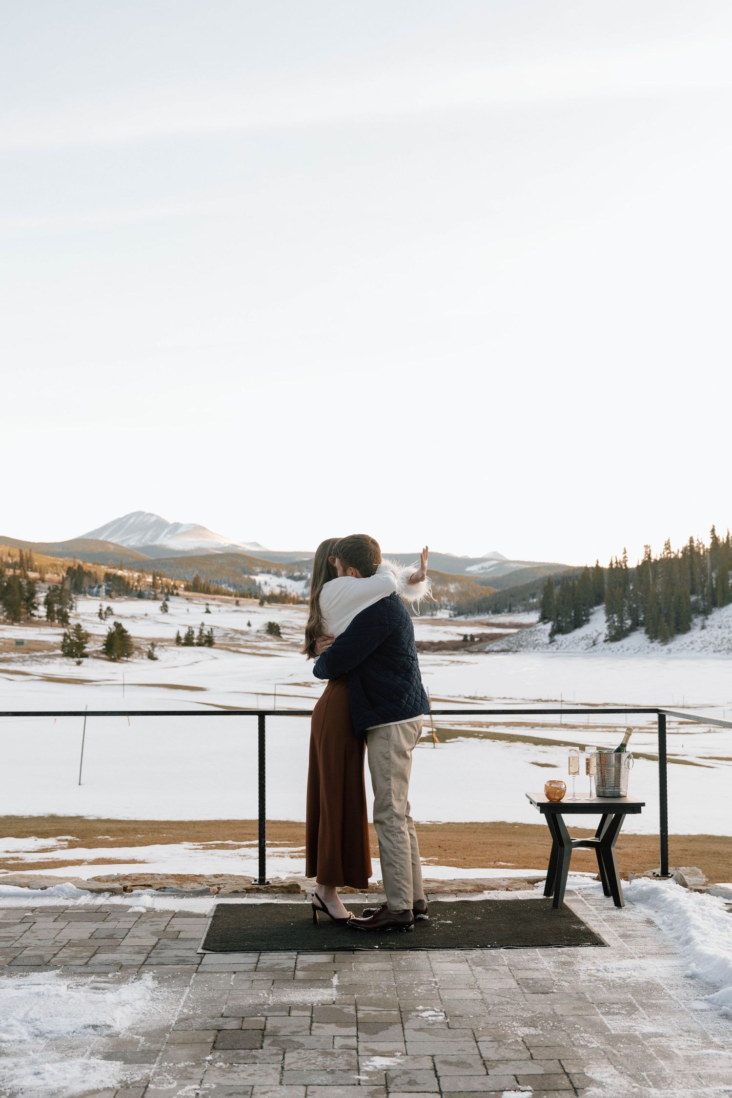 Newly engaged couple hugging on deck with snow-capped mountains in background.