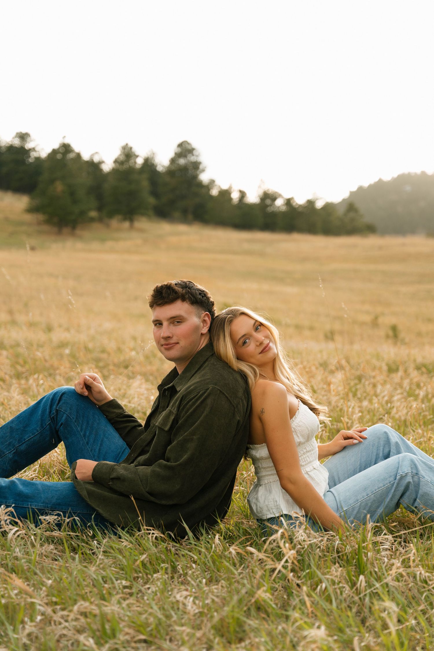 Couple sitting back-to-back in a golden field, smiling, with trees and soft sunset light in the background.