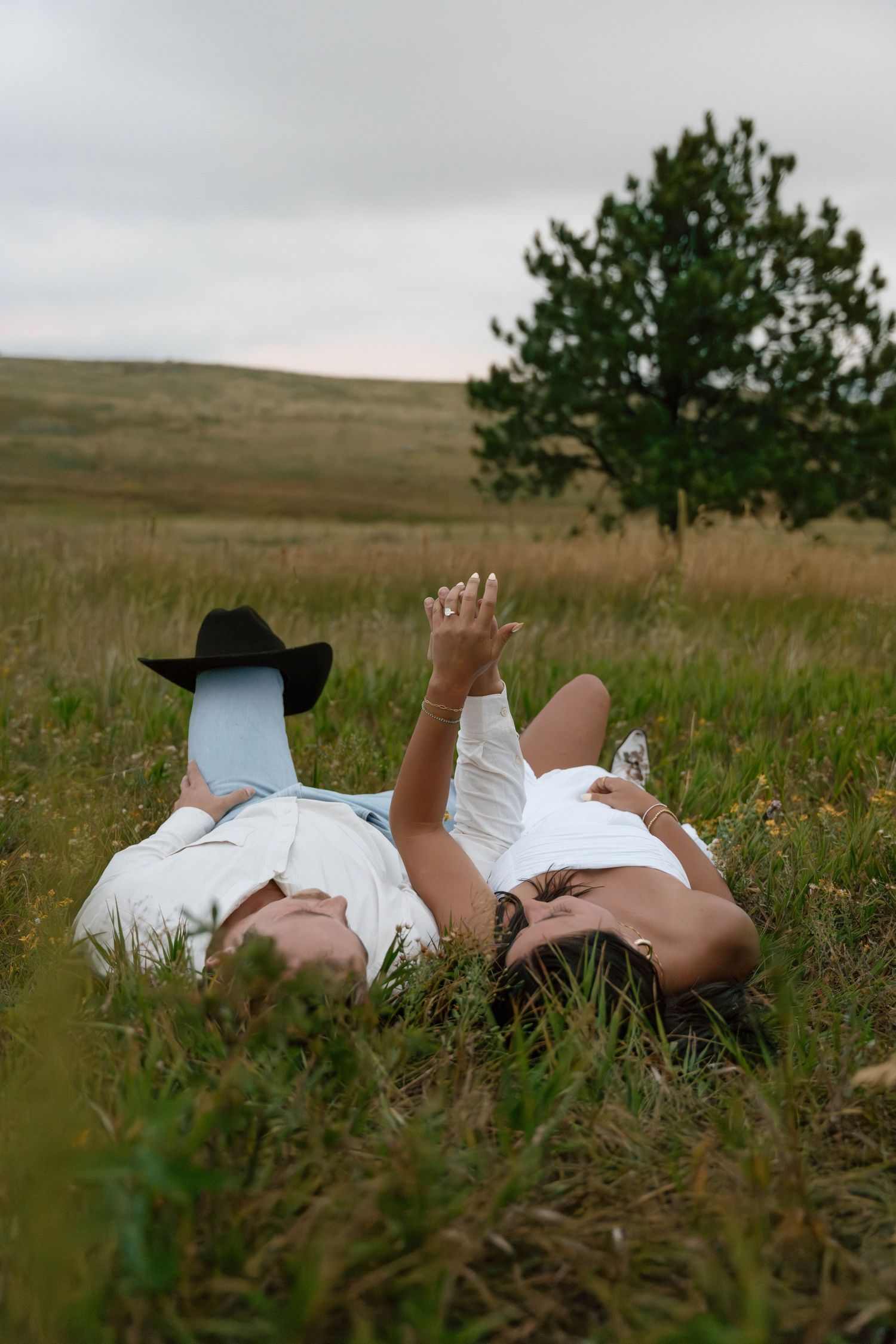 Couple lying in tall grass wearing western attire against mountain backdrop.
