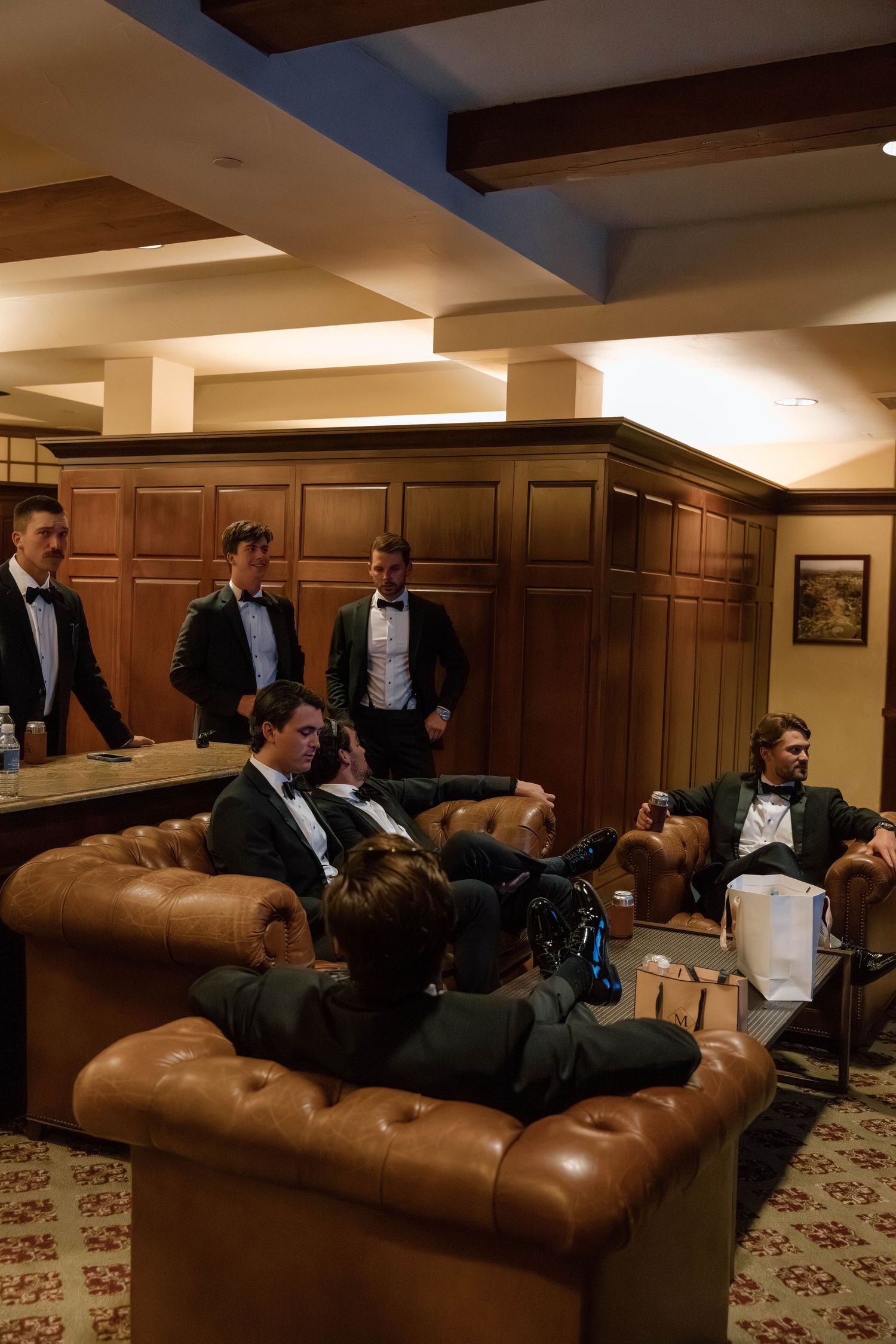 Groomsmen relaxing in leather chairs in a wood-paneled room with exposed ceiling beams.