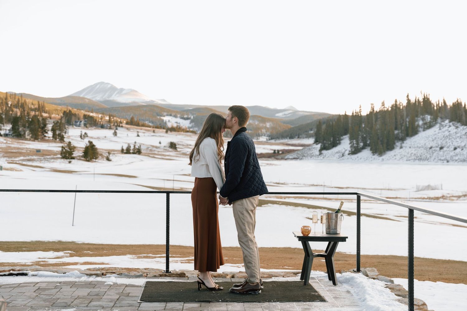 Romantic moment on snow-covered deck overlooking mountain landscape at sunset.