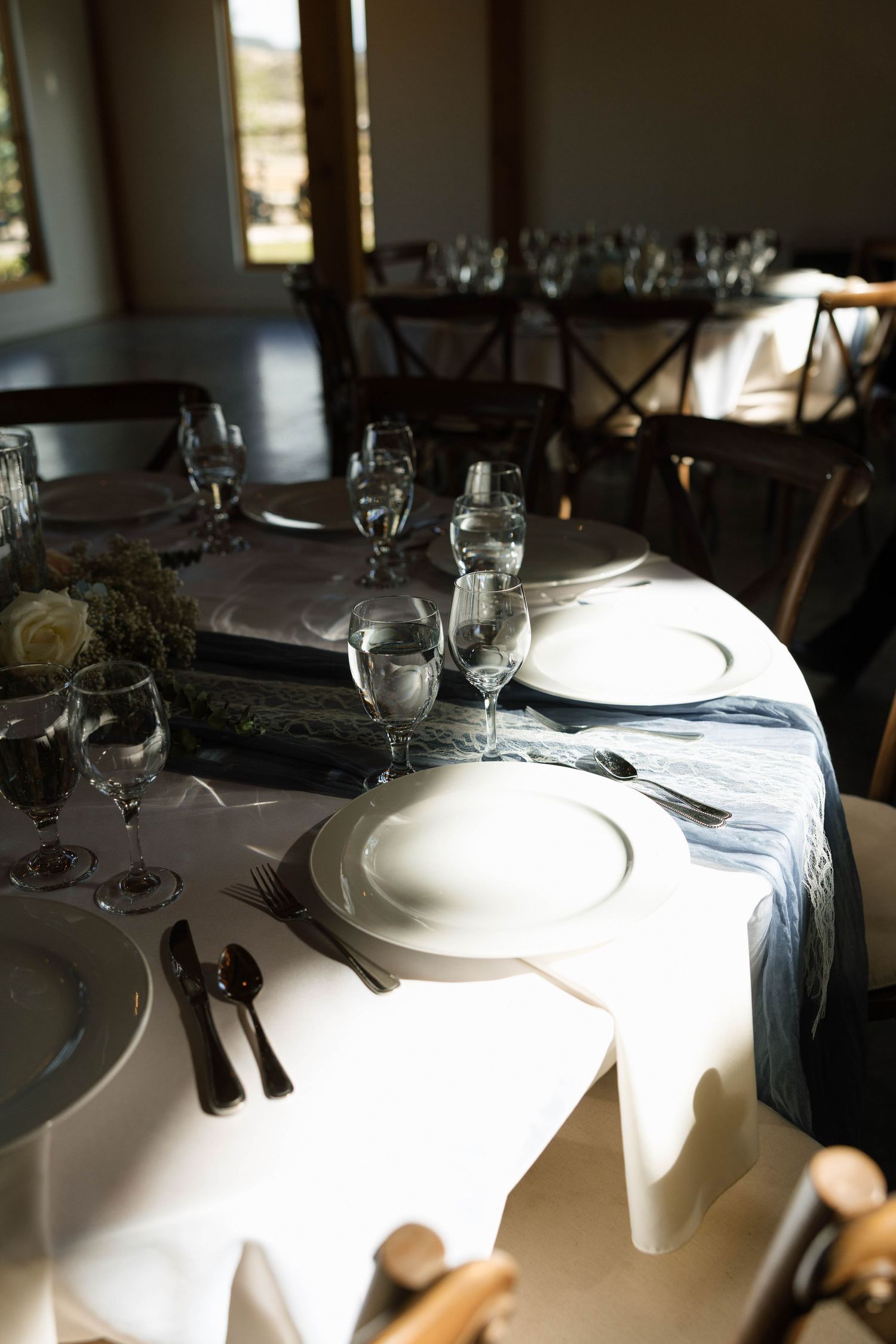 Elegant wedding reception table setting with white plates, crystal glasses, and blue linen runner in sunlit venue.