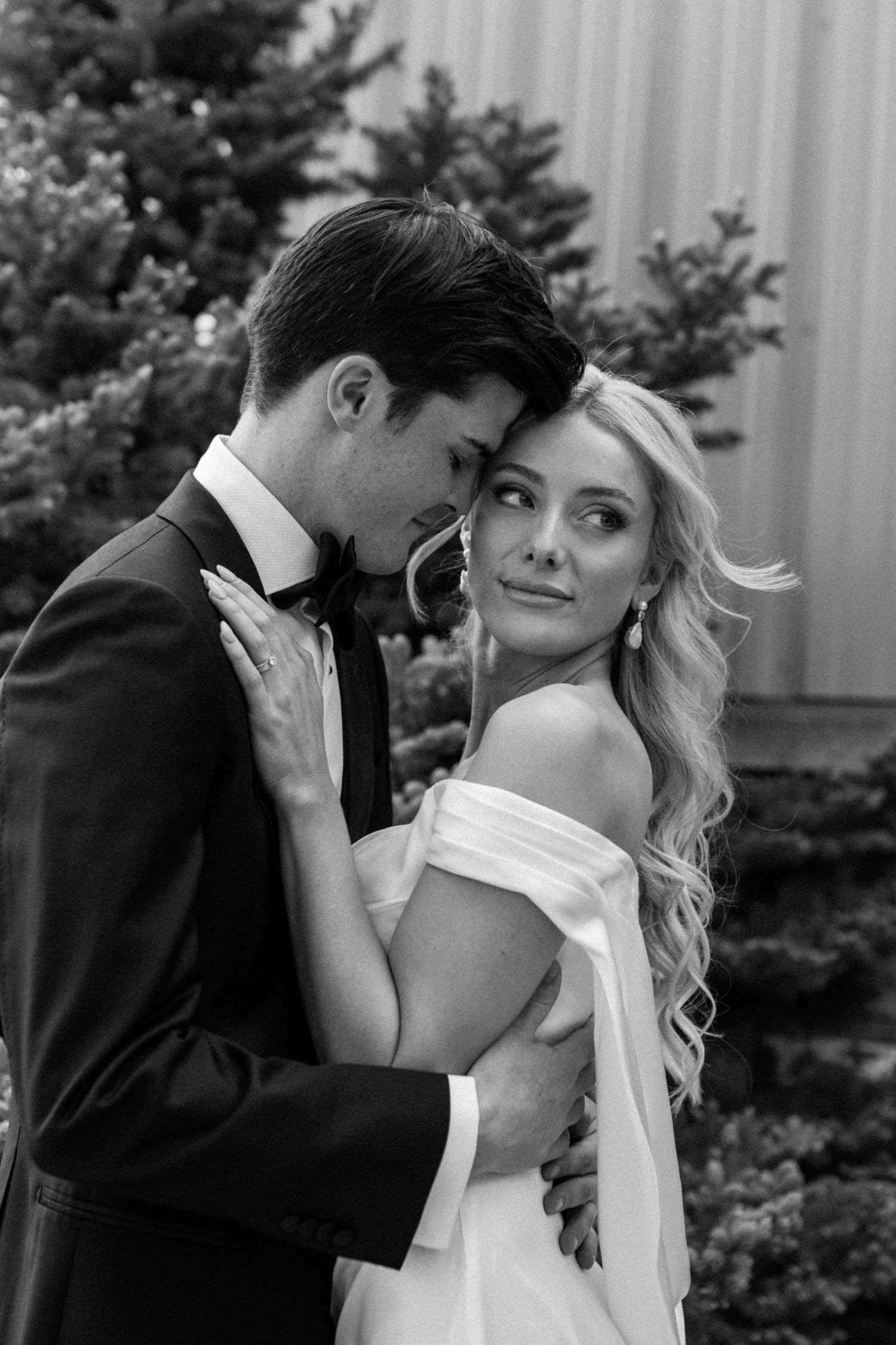 Black and white wedding photo of bride and groom embracing tenderly outdoors near evergreen trees.