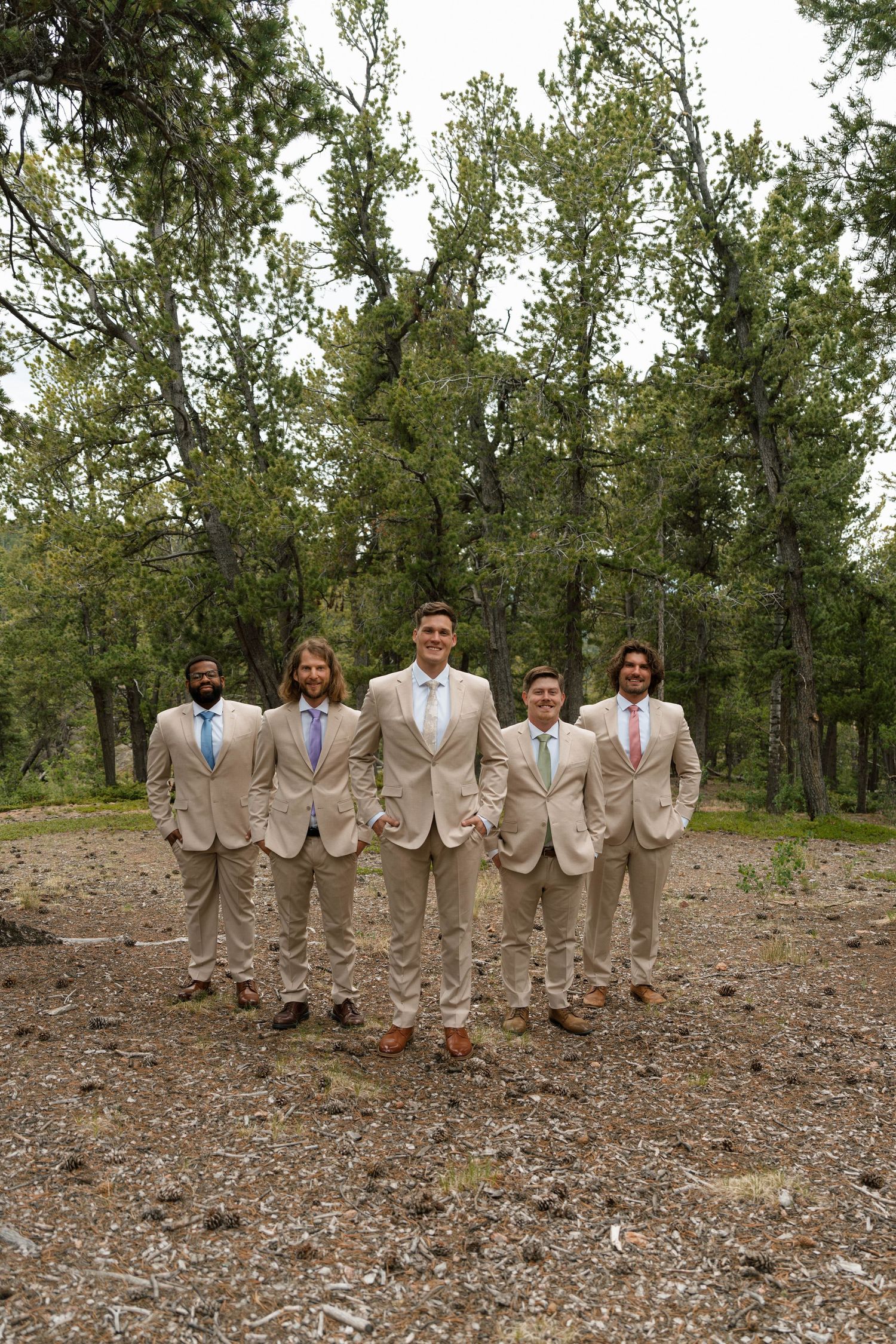 Groom and groomsmen in tan suits posing together in forest setting before wedding.