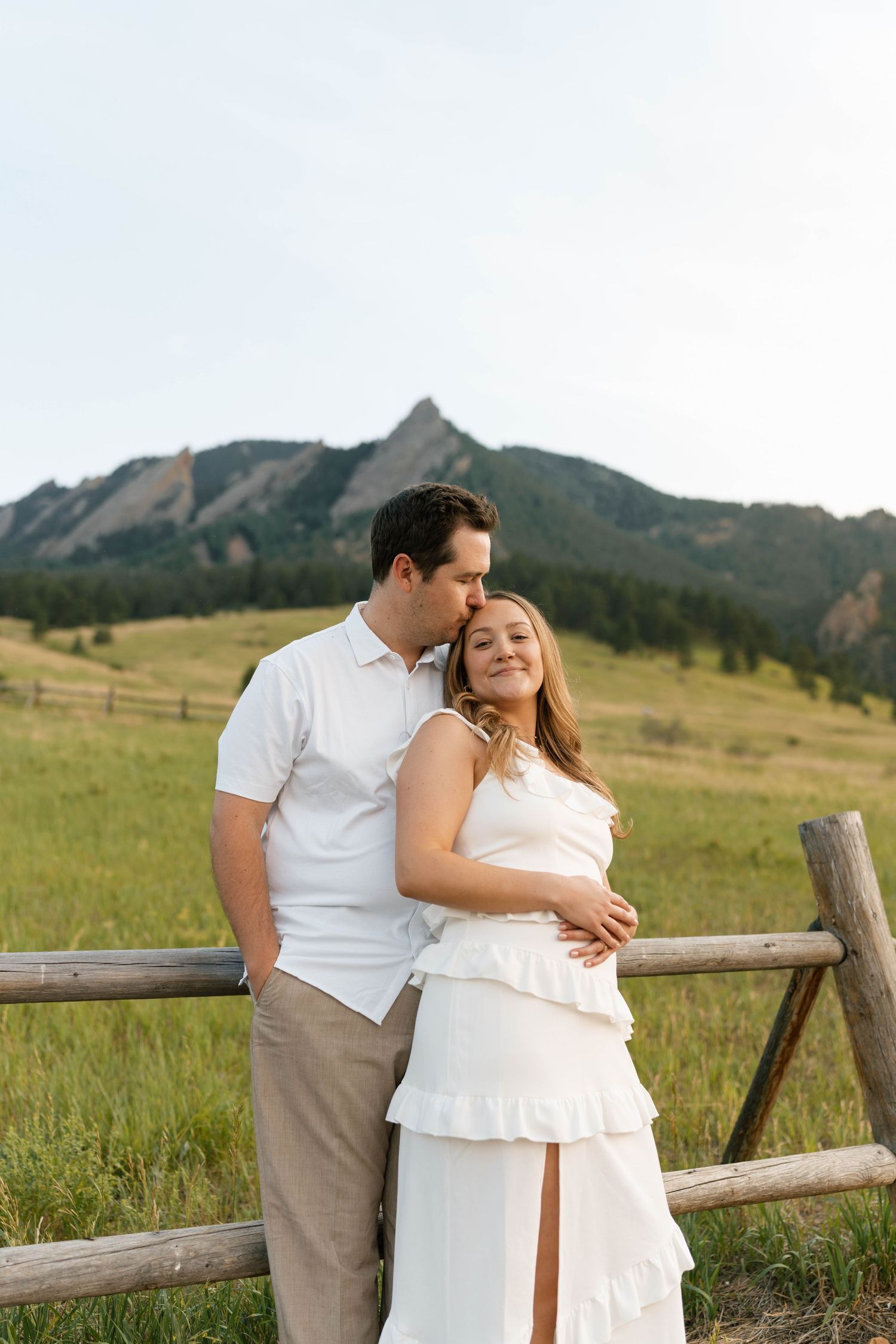 Romantic couple in white outfits embracing by a wooden fence with the Flatirons mountains behind them.