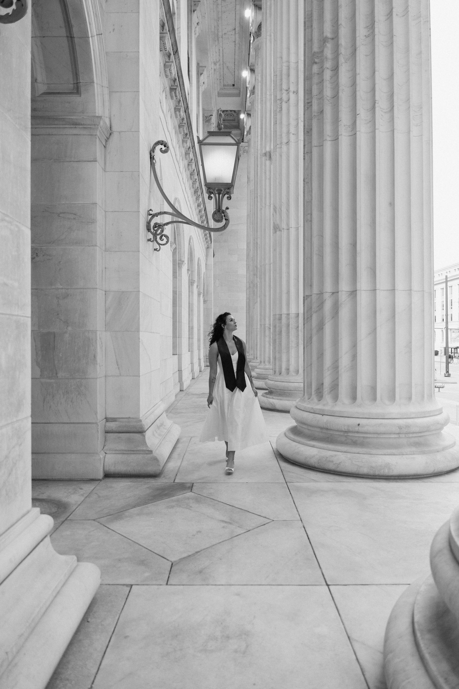 Woman in white skirt walks among grand marble columns of a classical building in black and white photo.