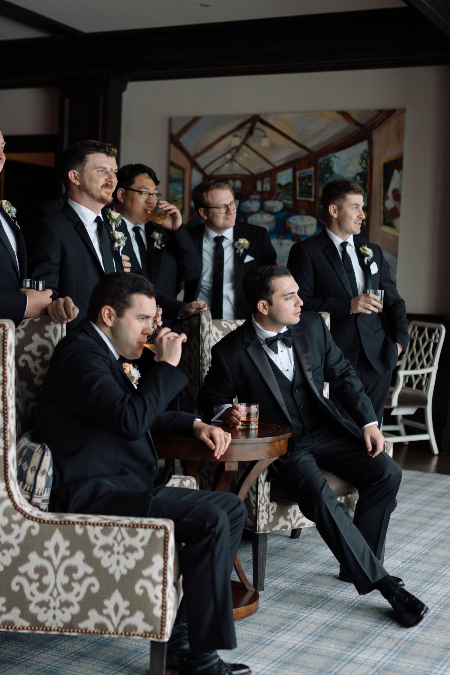 Groomsmen in black tuxedos with boutonnieres relaxing in elegant lounge, some holding drinks before wedding ceremony.