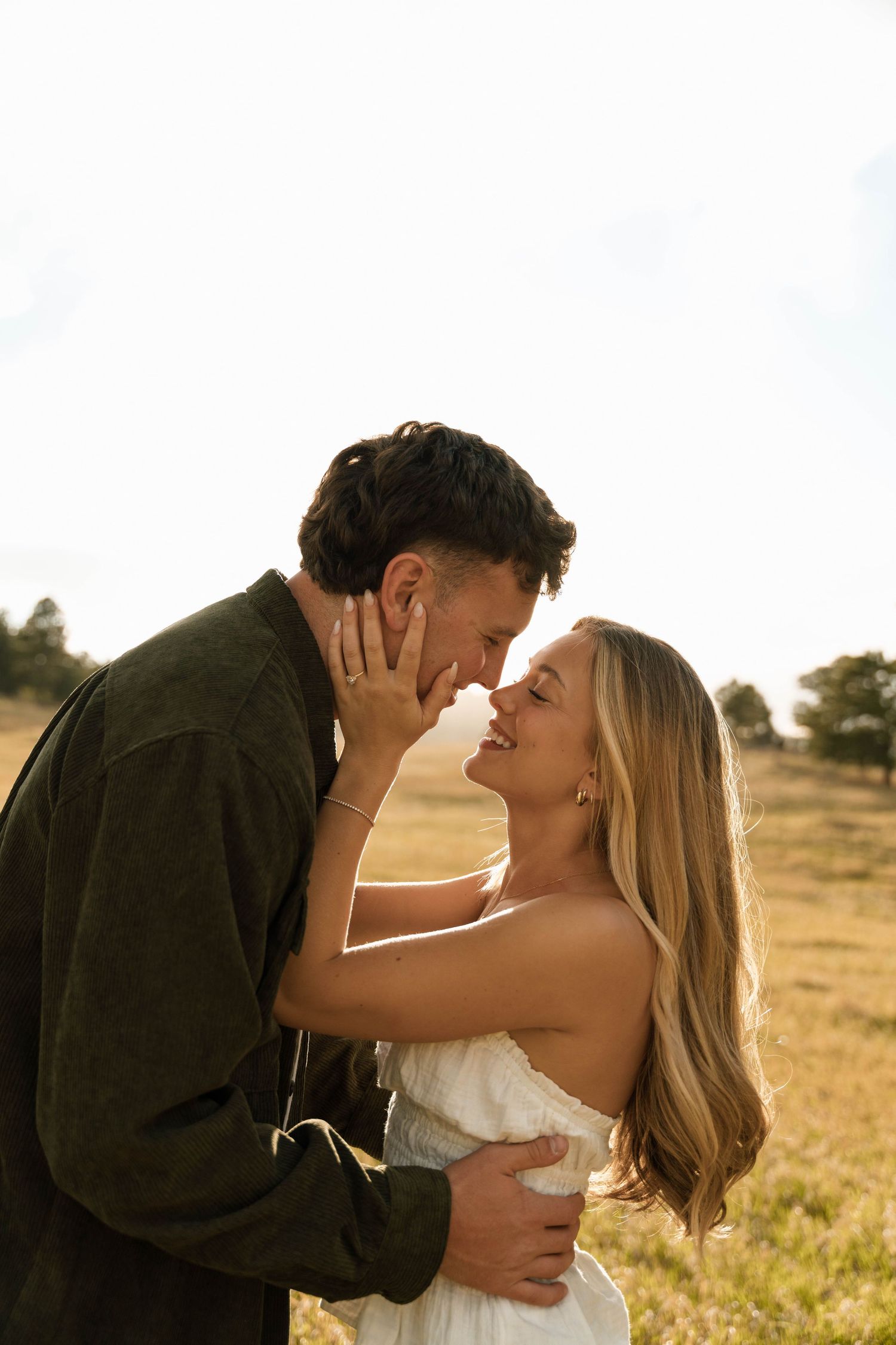 A couple shares a romantic almost-kiss moment in a sunlit golden field, woman in white dress smiling up at man in green jacket.