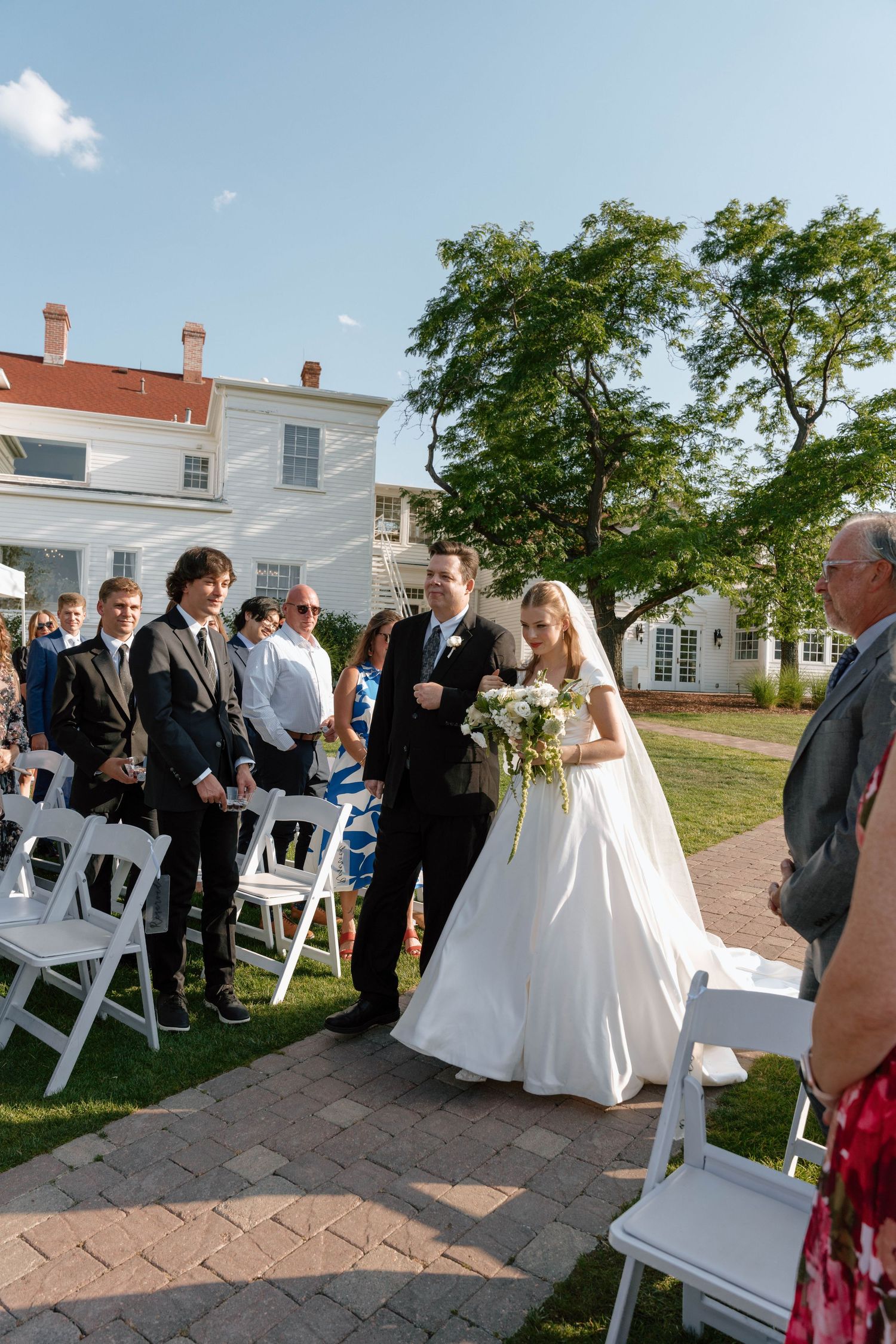 A wedding ceremony procession on a brick pathway with a white colonial house and green lawn.