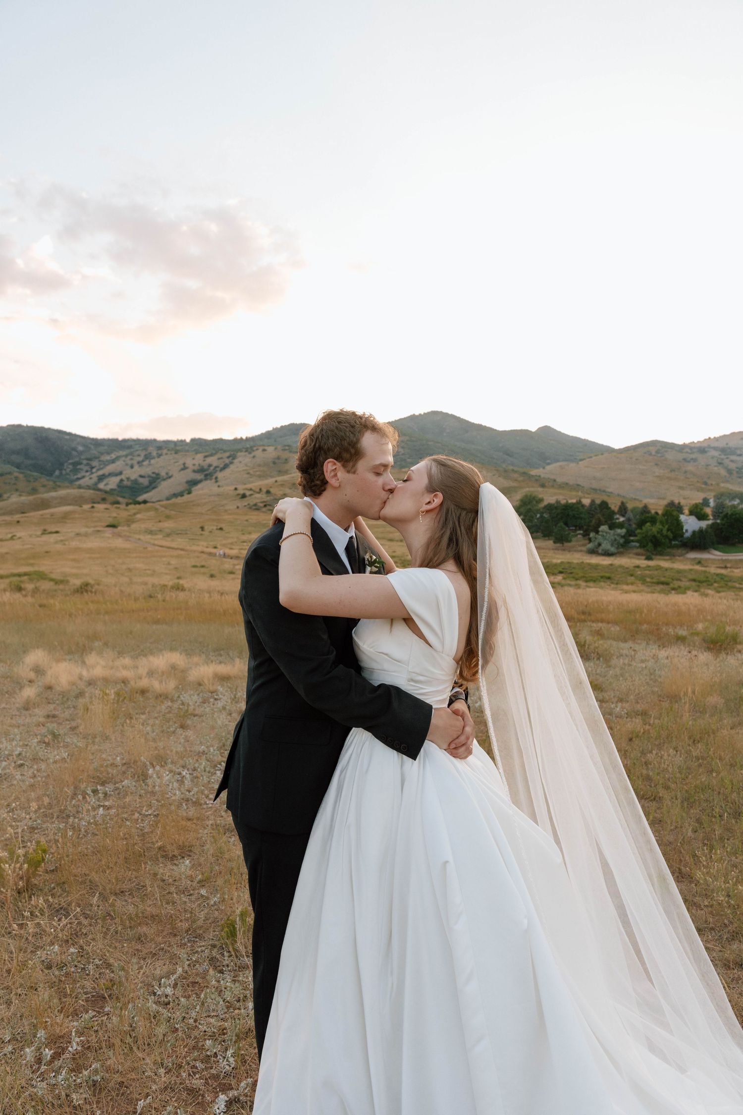 Newlyweds share intimate moment in golden field with mountain backdrop at sunset.
