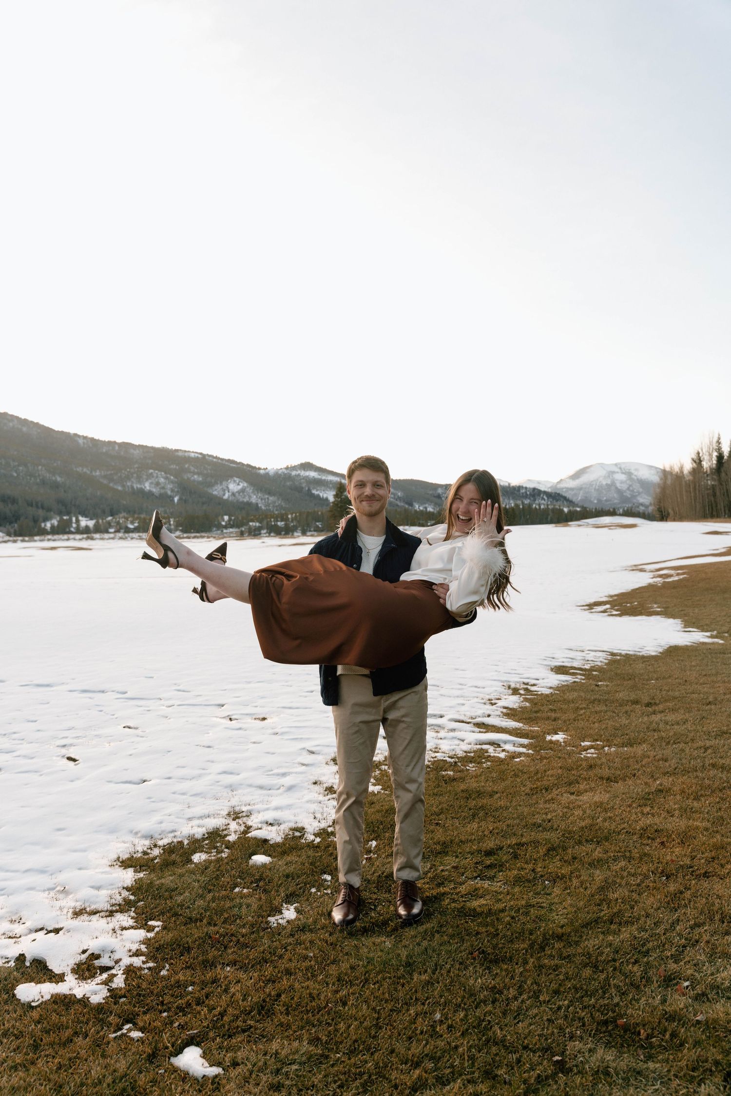 Playful couple posing with arms outstretched on snow-covered ground near mountains.