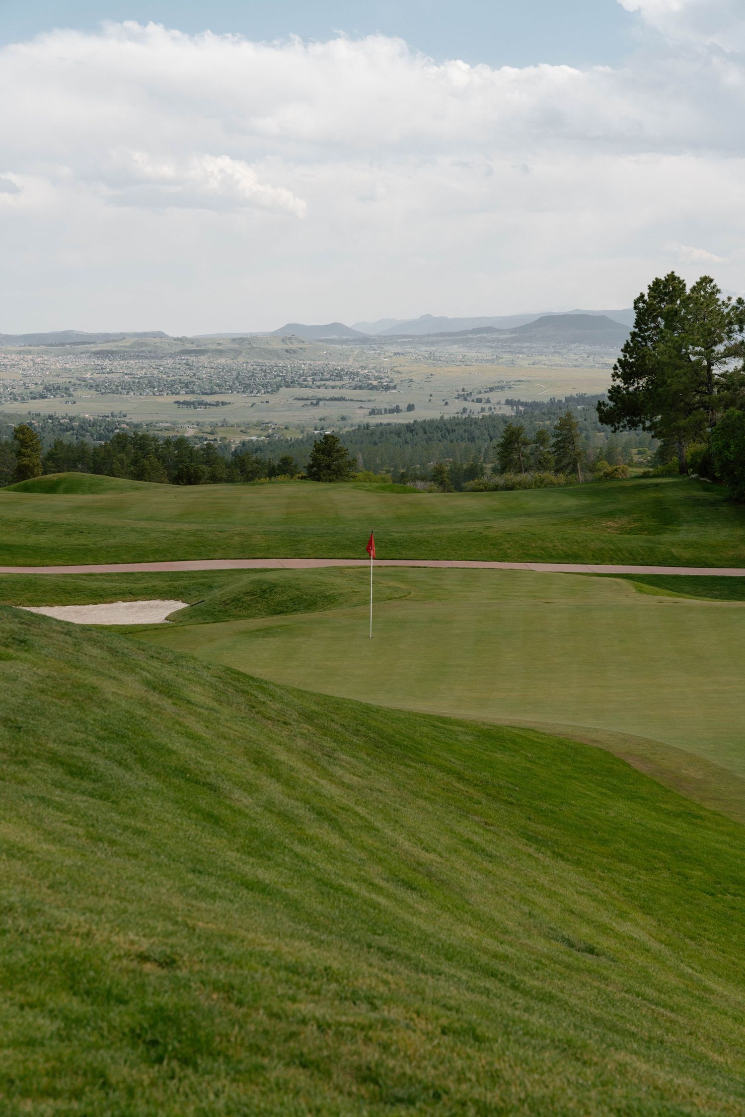 Rolling green golf course with mountain views and a flagged putting green in the distance.