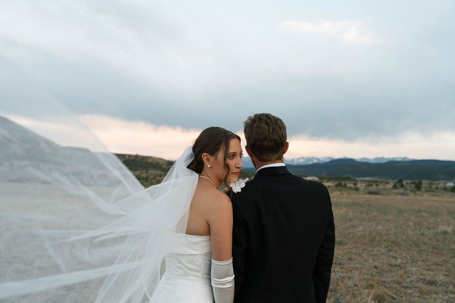 Bride and groom share an intimate moment outdoors with veil blowing in wind against mountain backdrop.