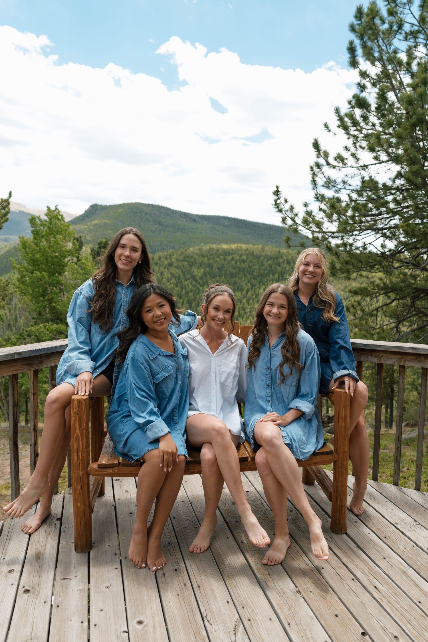 Five women in blue robes sit on a wooden deck with mountain and forest views, smiling together outdoors.