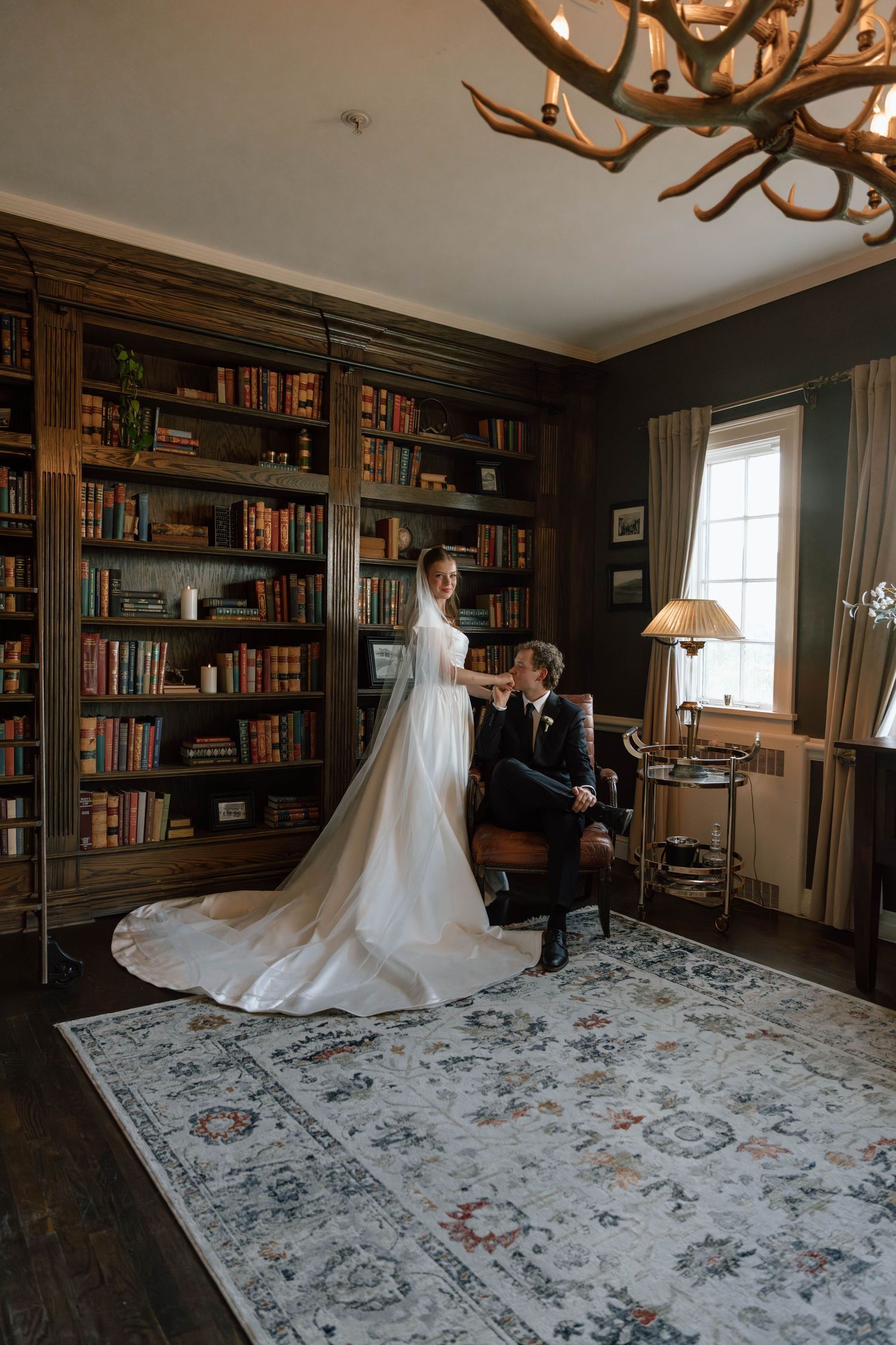 Wedding dress displayed in an elegant library room with floor-to-ceiling bookshelves and antler chandelier.