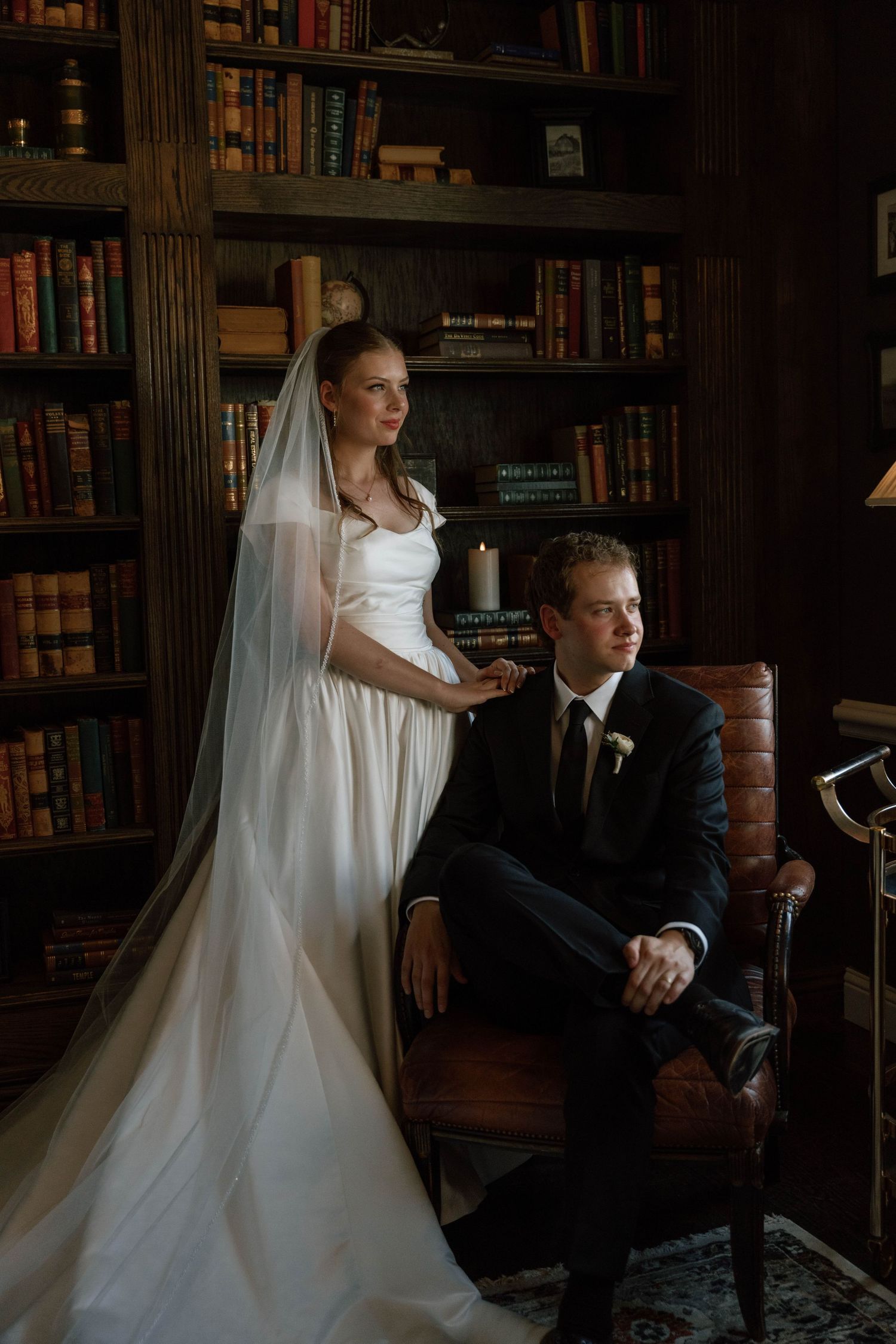 Moody wedding portrait in dark wood-paneled library with floor-to-ceiling bookshelves.