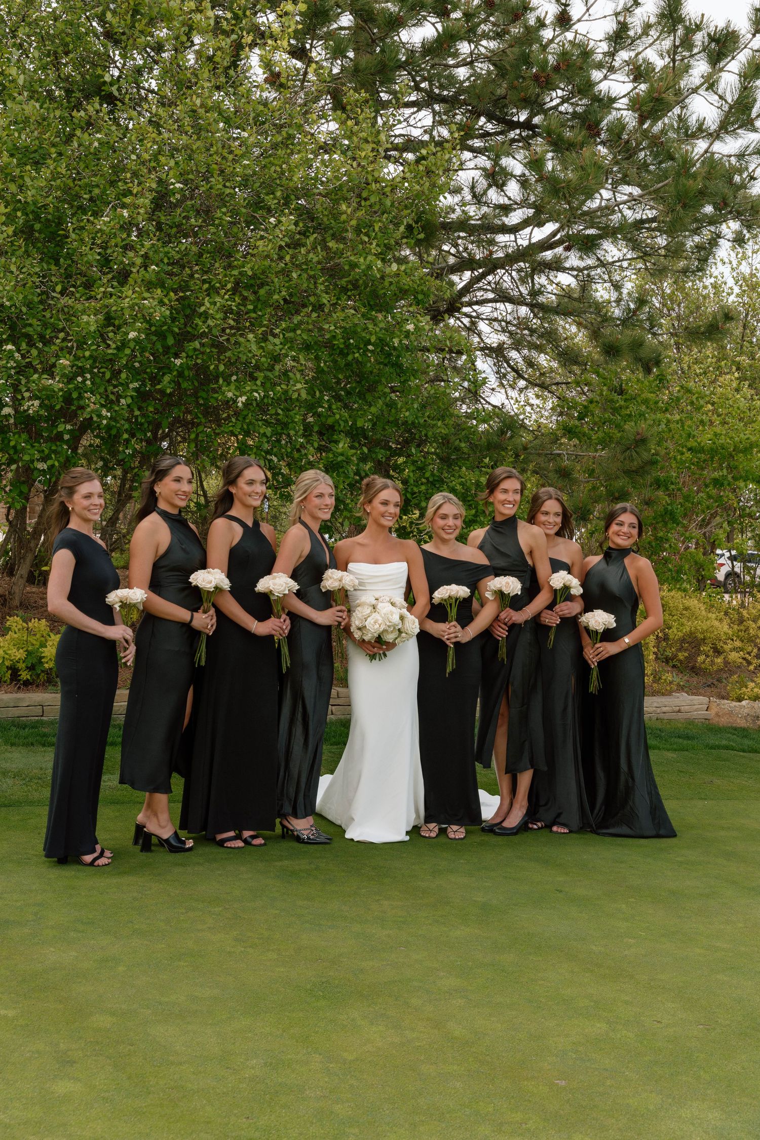 A wedding party in black and white attire posing on a manicured lawn with trees in the background.