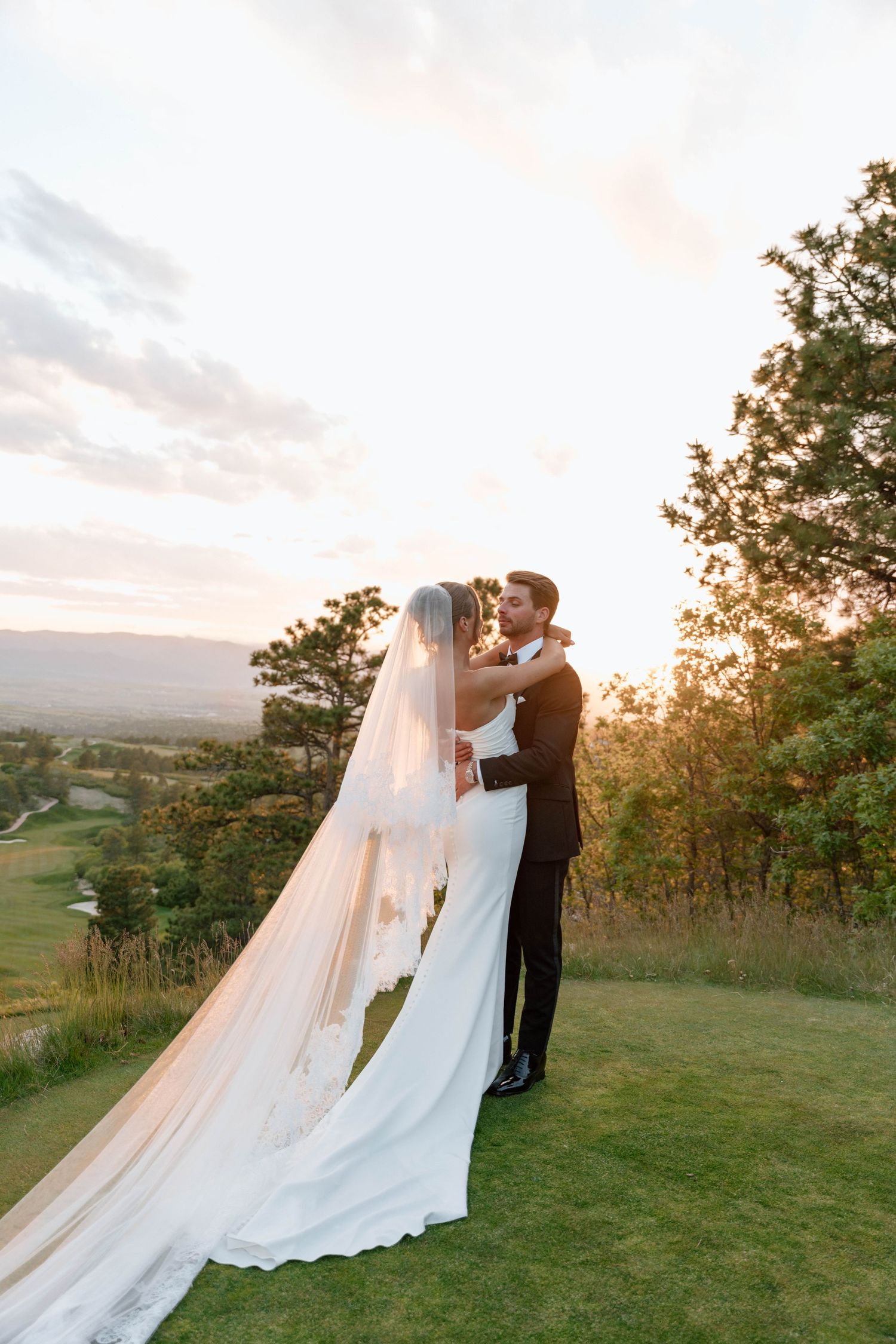 Wedding couple poses on hilltop with dramatic flowing veil and dress against sunset landscape.