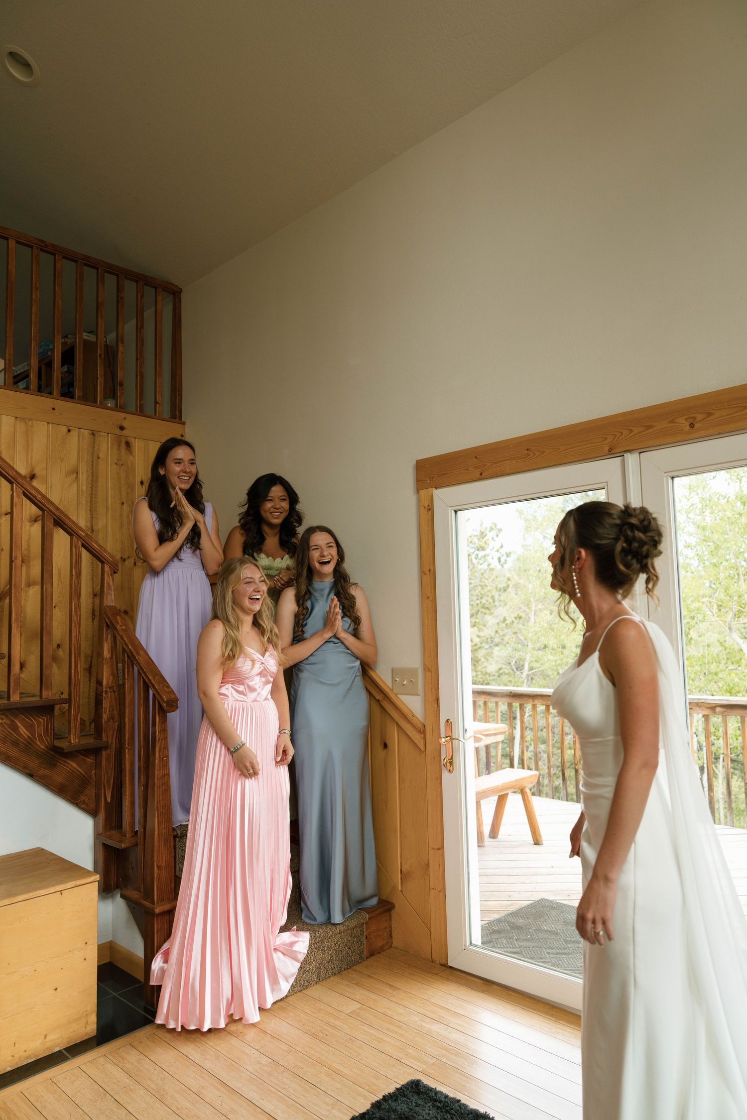 Bride reveals wedding dress to excited bridesmaids in colorful gowns on a staircase inside a rustic cabin.