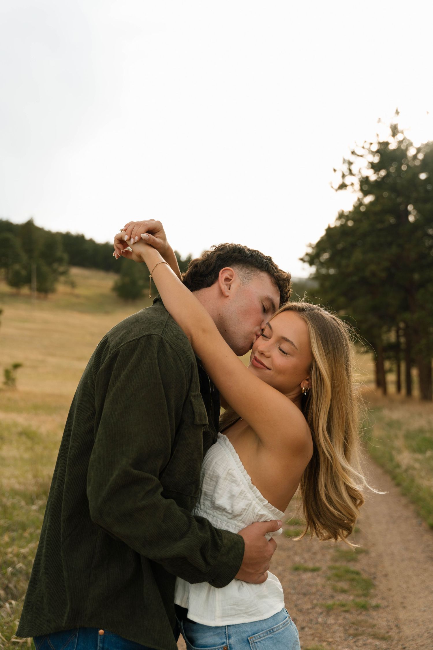 A couple shares a tender kiss outdoors on a scenic path, the man in green embracing a blonde woman in white.