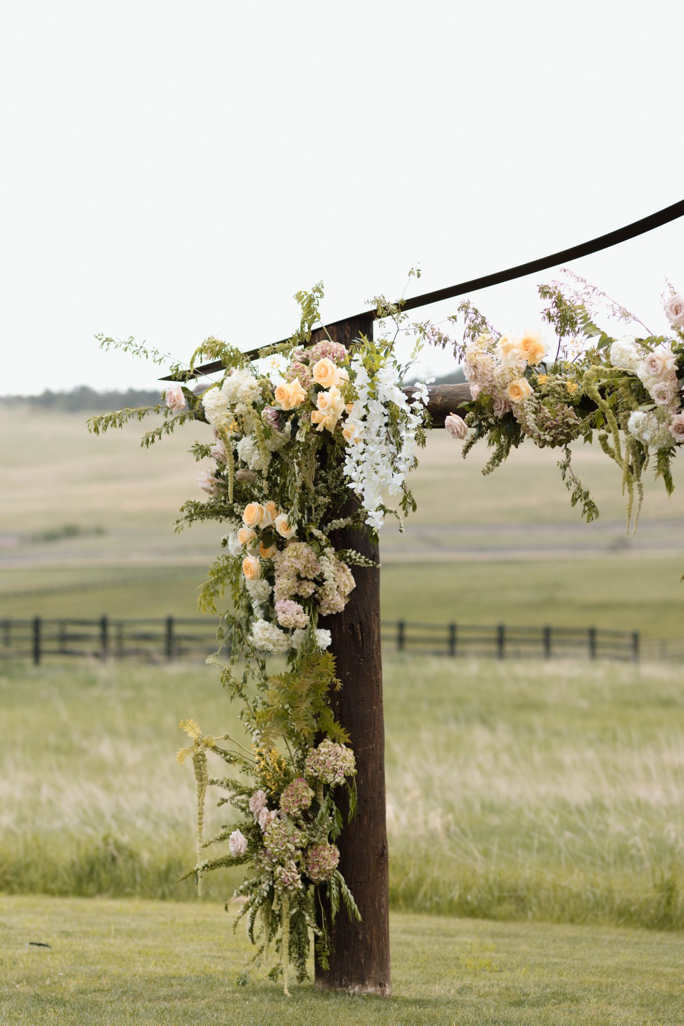 Rustic wooden wedding arch adorned with lush greenery and soft peach, white floral arrangements in an open field.