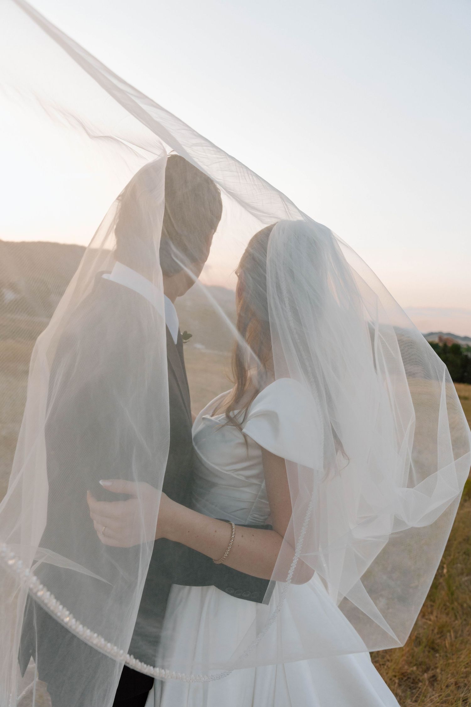 Bride and groom sharing a romantic moment under a flowing white veil at sunset in an outdoor field.