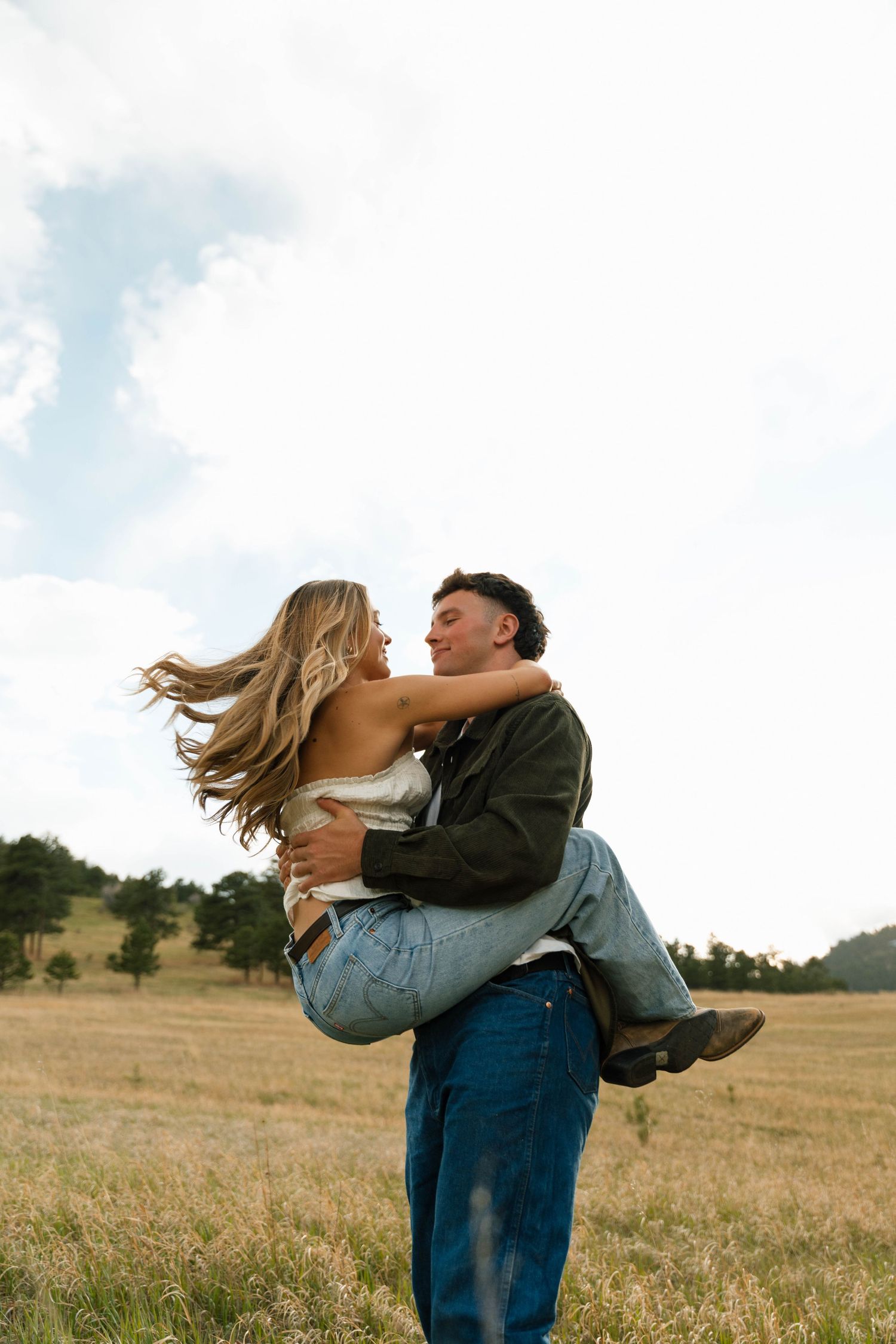 A man lifts and spins a woman in a golden field, both smiling joyfully under a cloudy sky.