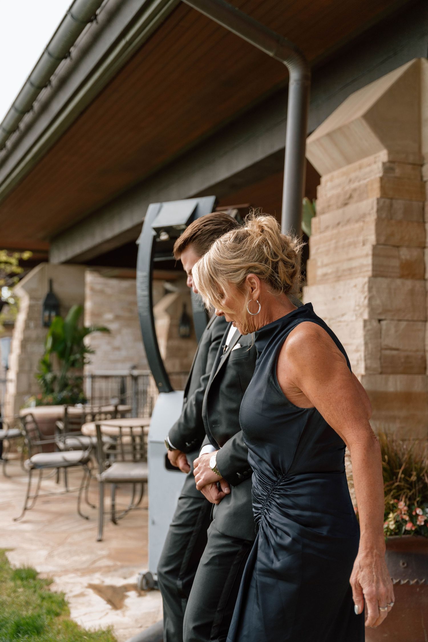 Wedding guests in formal attire gathering under a covered stone patio area.