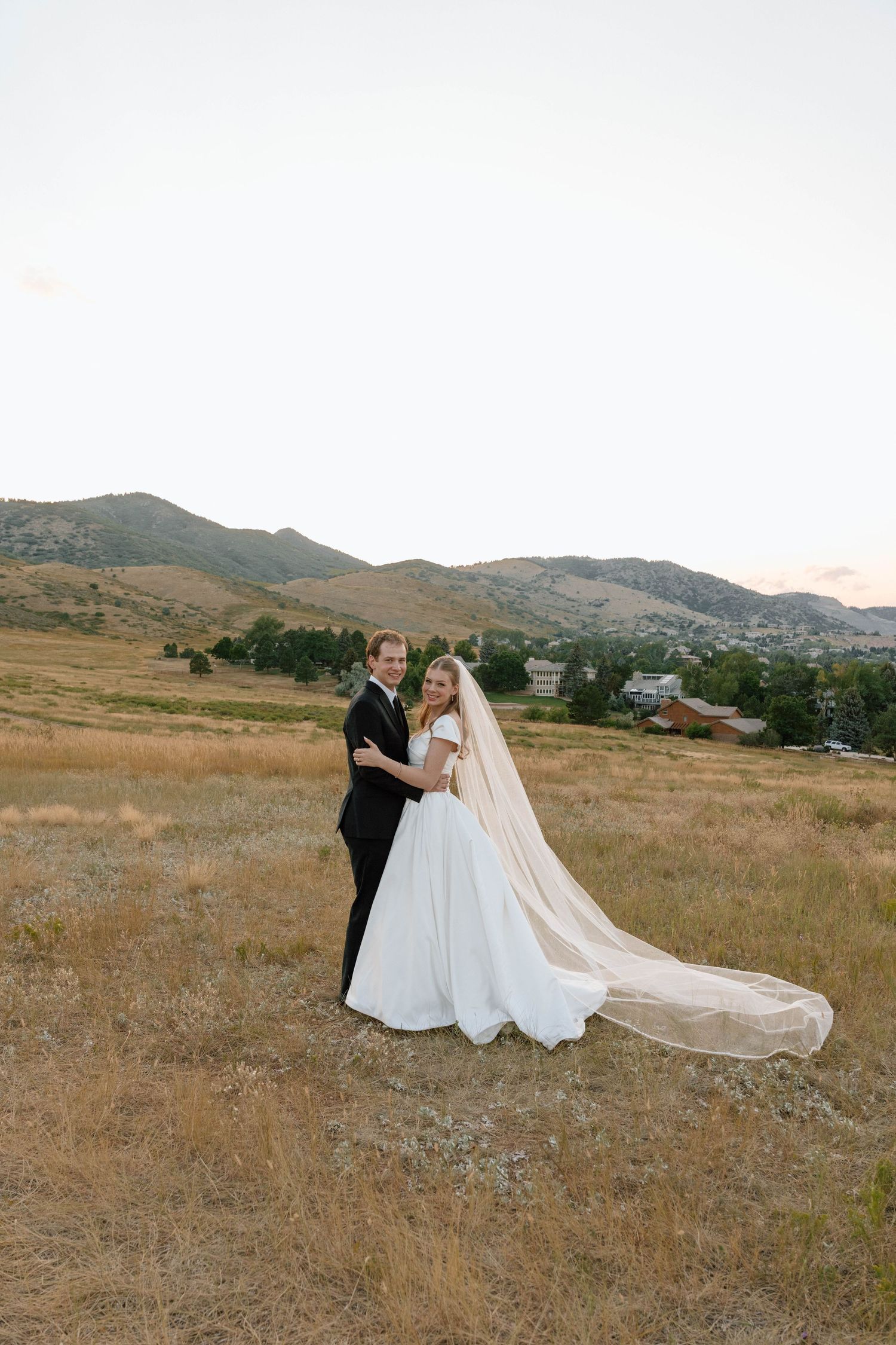 Wedding couple poses in mountain field with dramatic long veil trailing behind them.