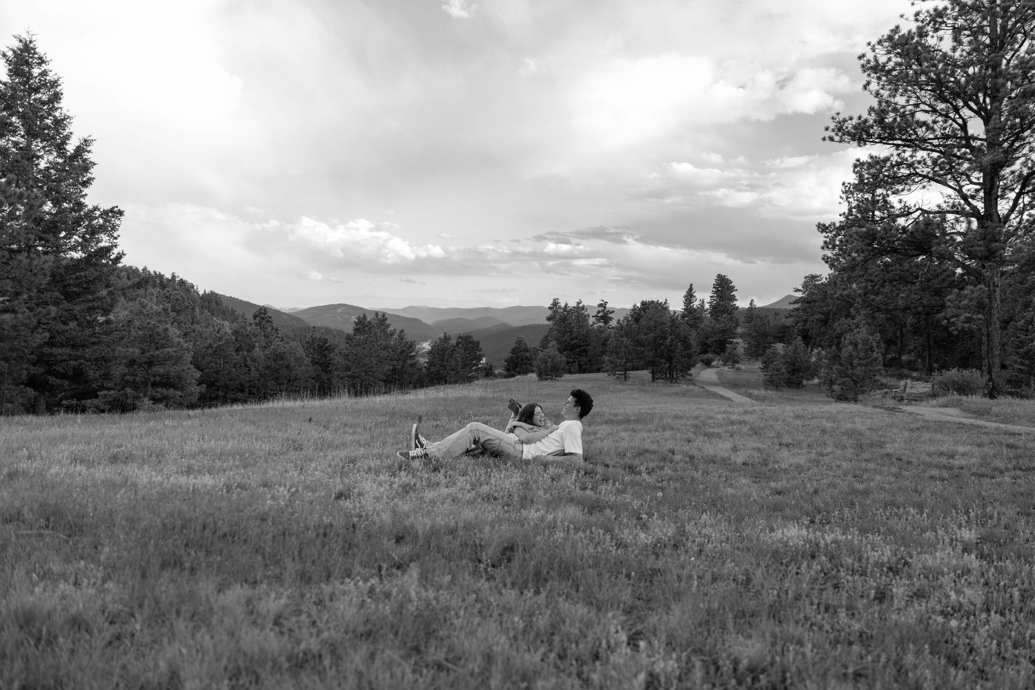 Black and white photo of a person relaxing in a vast meadow surrounded by trees and rolling hills under a cloudy sky.