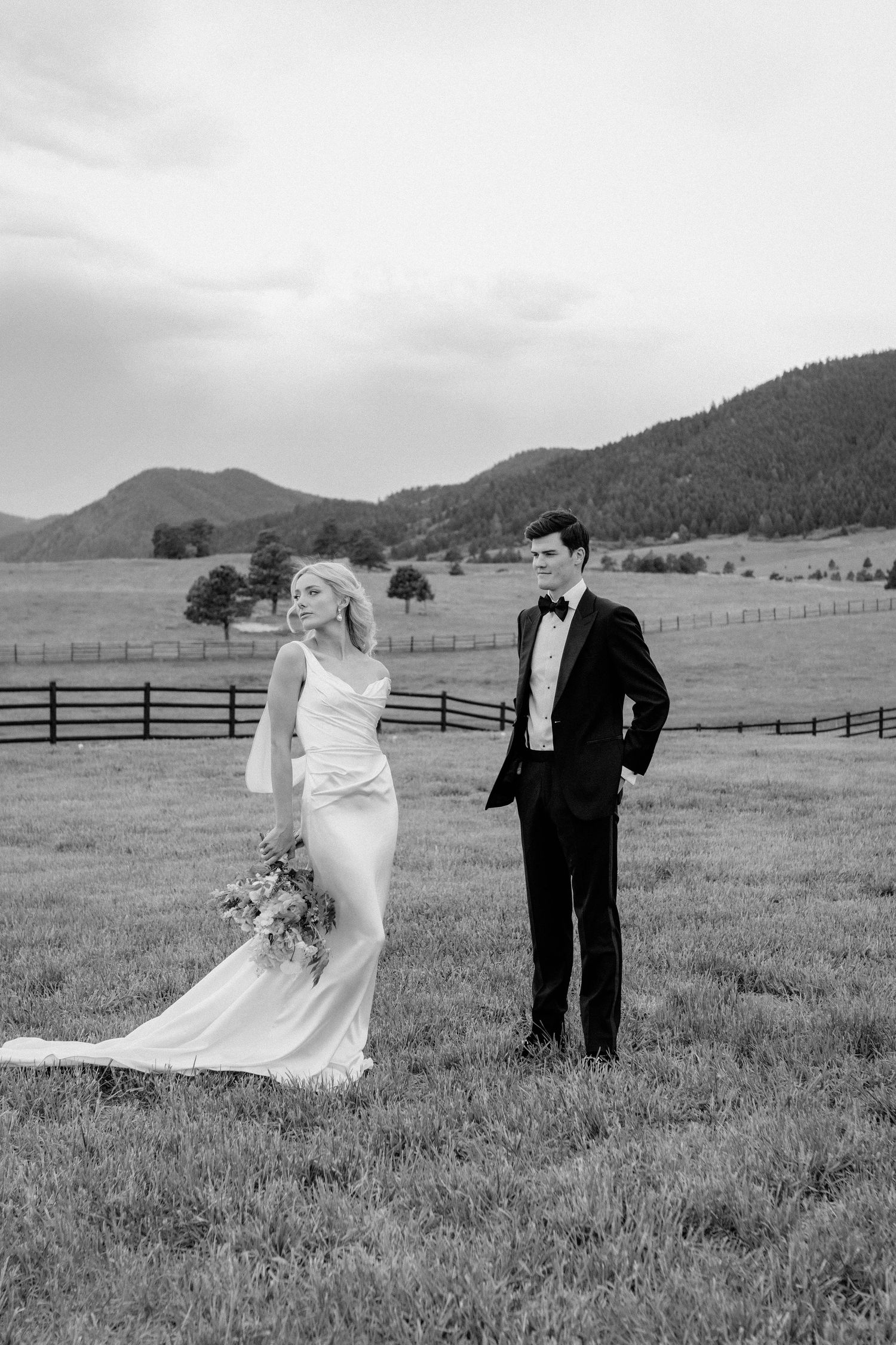 Bride and groom stand in scenic mountain meadow with fence in elegant black and white wedding photo.