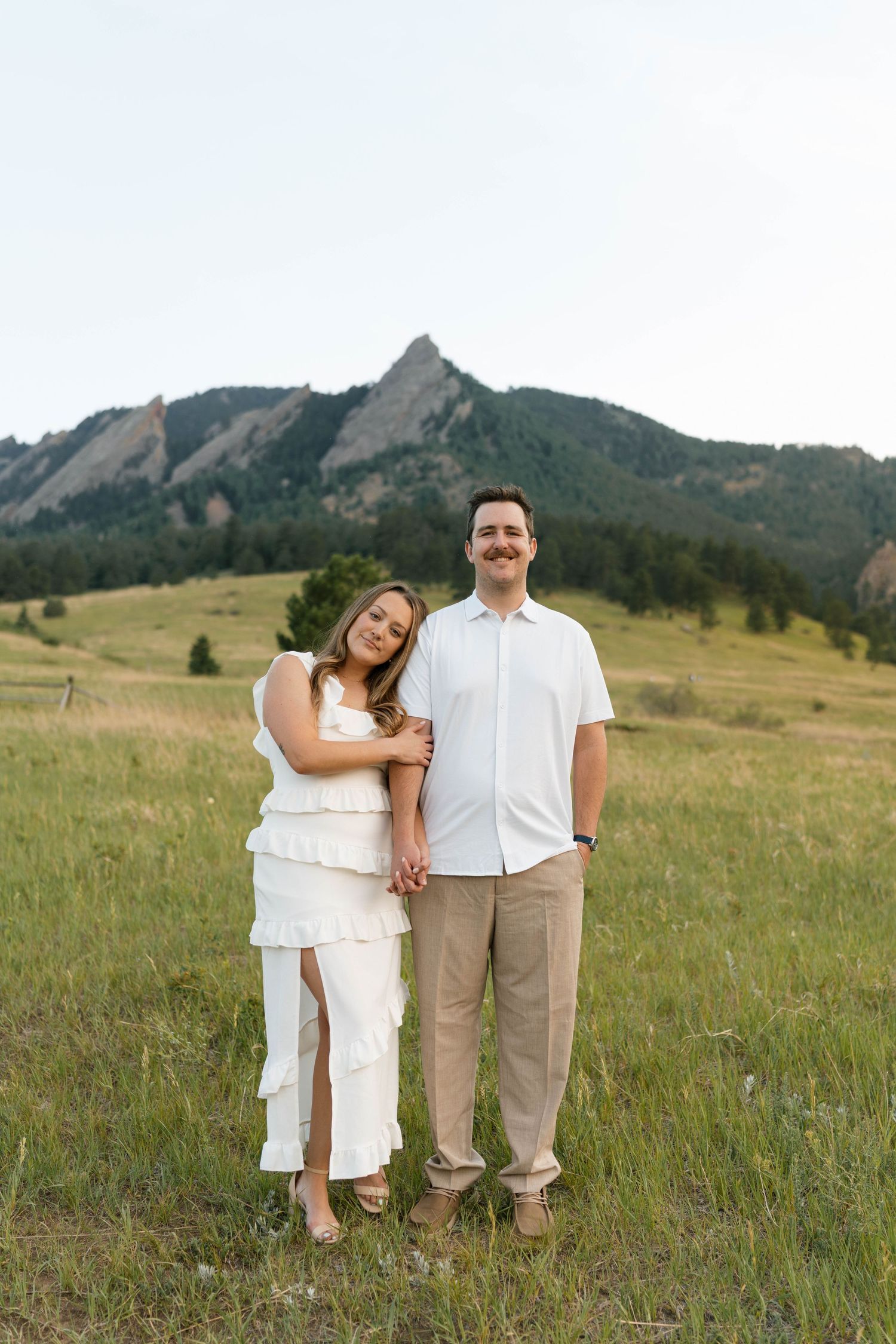 Couple posing in white and khaki outfits in a green meadow with the Flatirons mountains in Boulder, Colorado.
