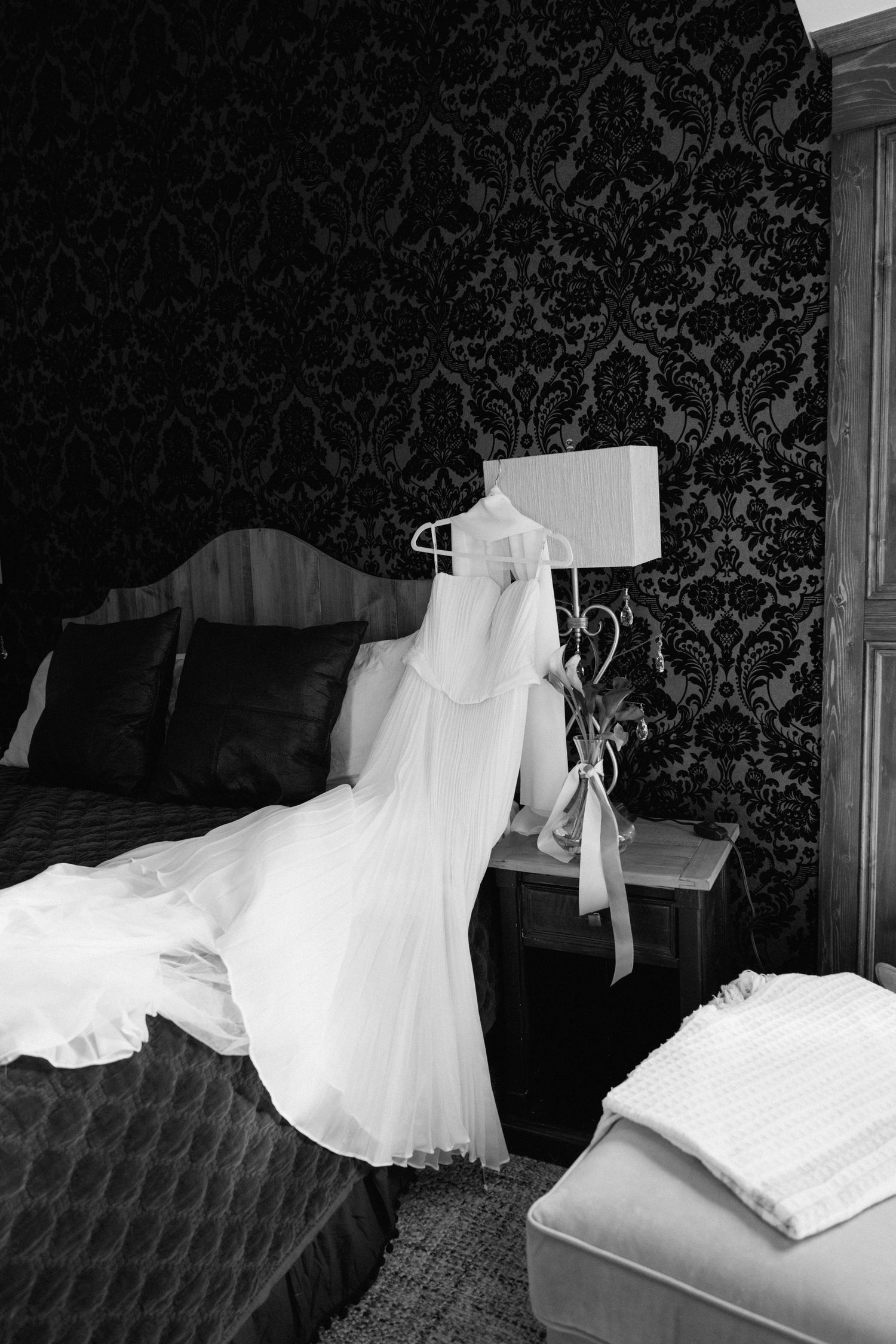 Black and white photo of a white wedding dress hanging on a lamp beside a bed in an elegant hotel room.