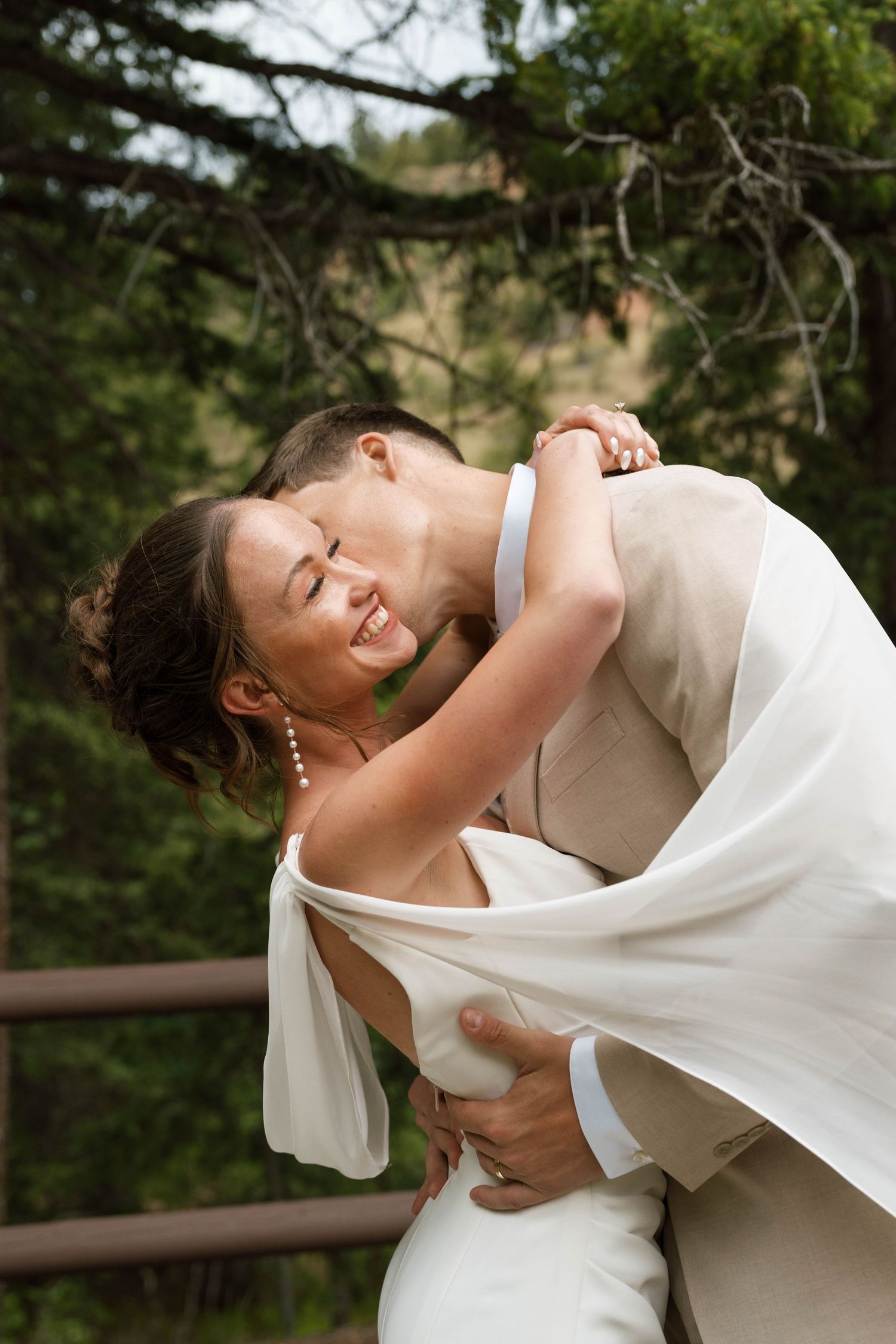 Groom dipping and kissing bride outdoors surrounded by pine trees after ceremony.