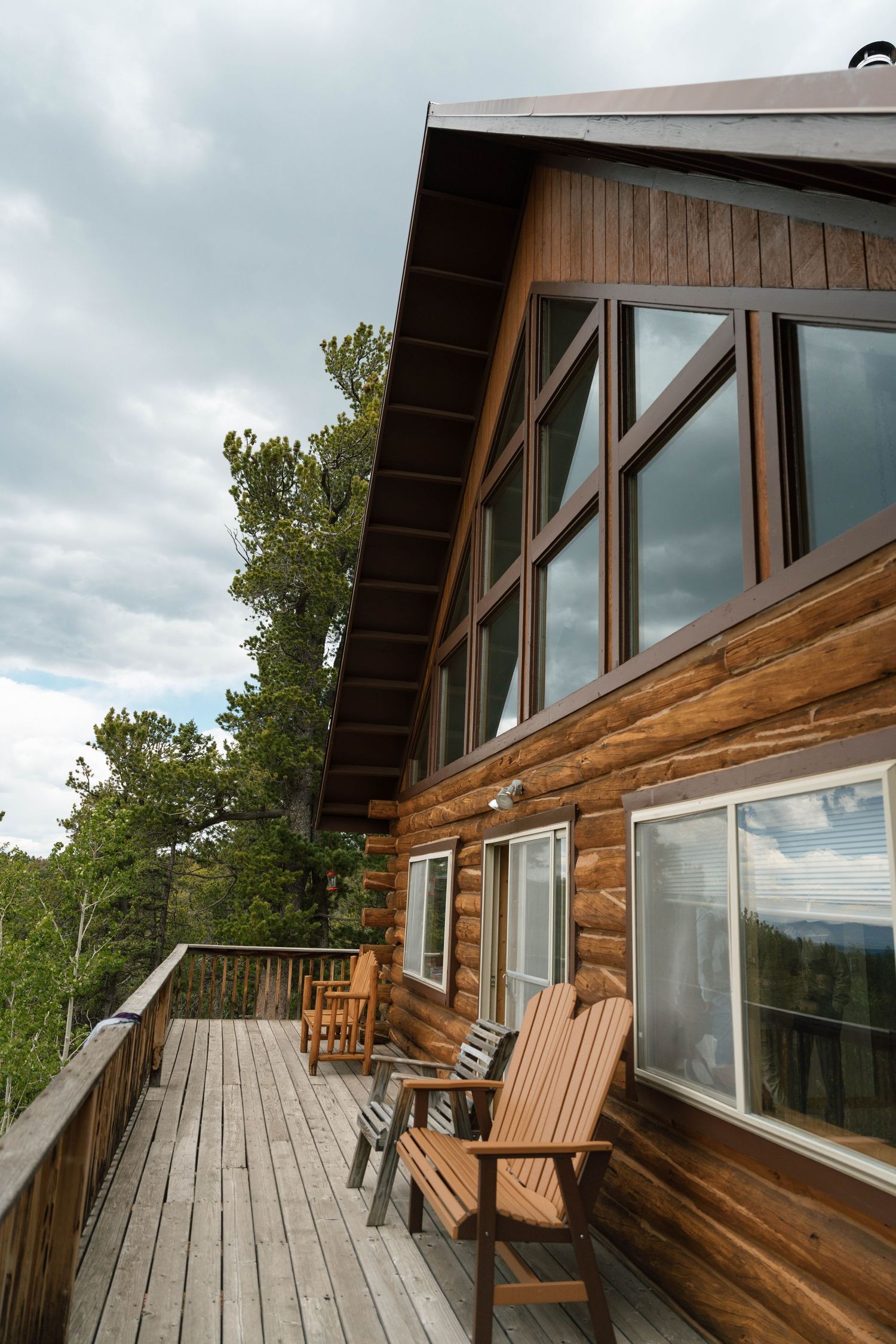 Rustic log cabin with large windows and wooden deck featuring Adirondack chairs under cloudy sky.