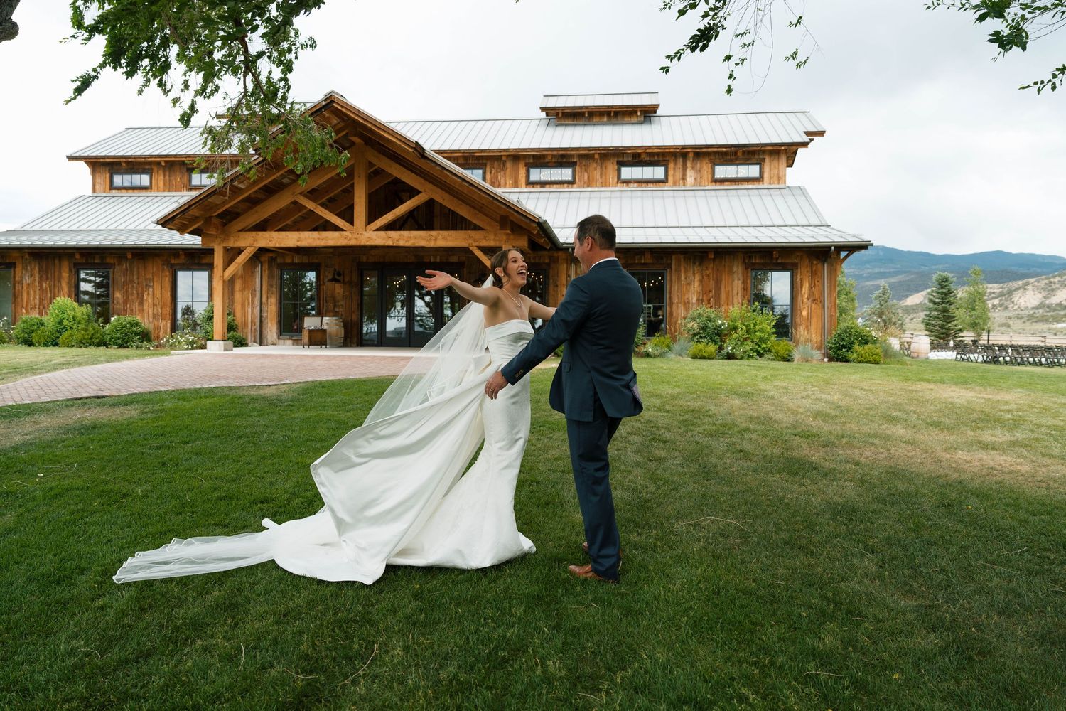 Bride and groom dancing on green lawn in front of rustic wooden wedding venue with mountain views.