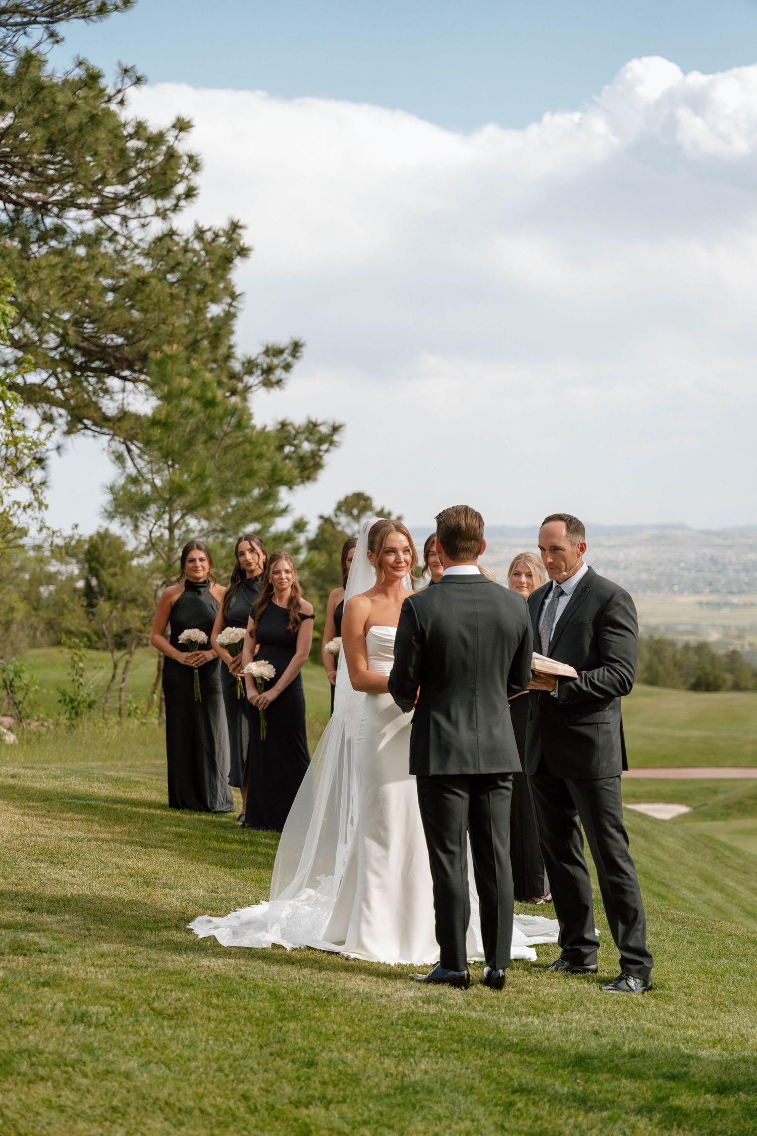 Outdoor wedding ceremony on rolling hills with attendants in black formal attire.