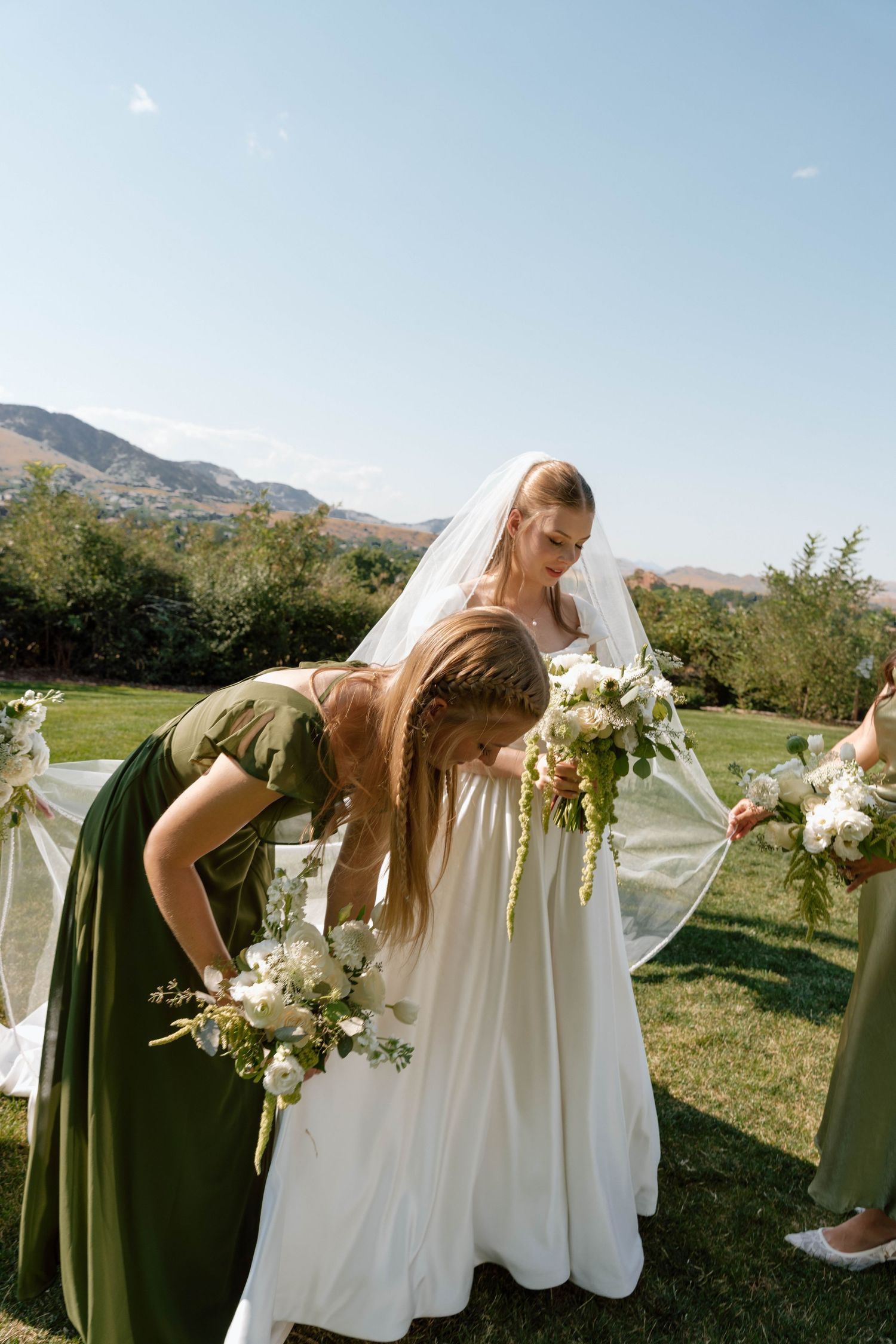 Wedding party members in green dresses arrange flowers outdoors with mountains visible.