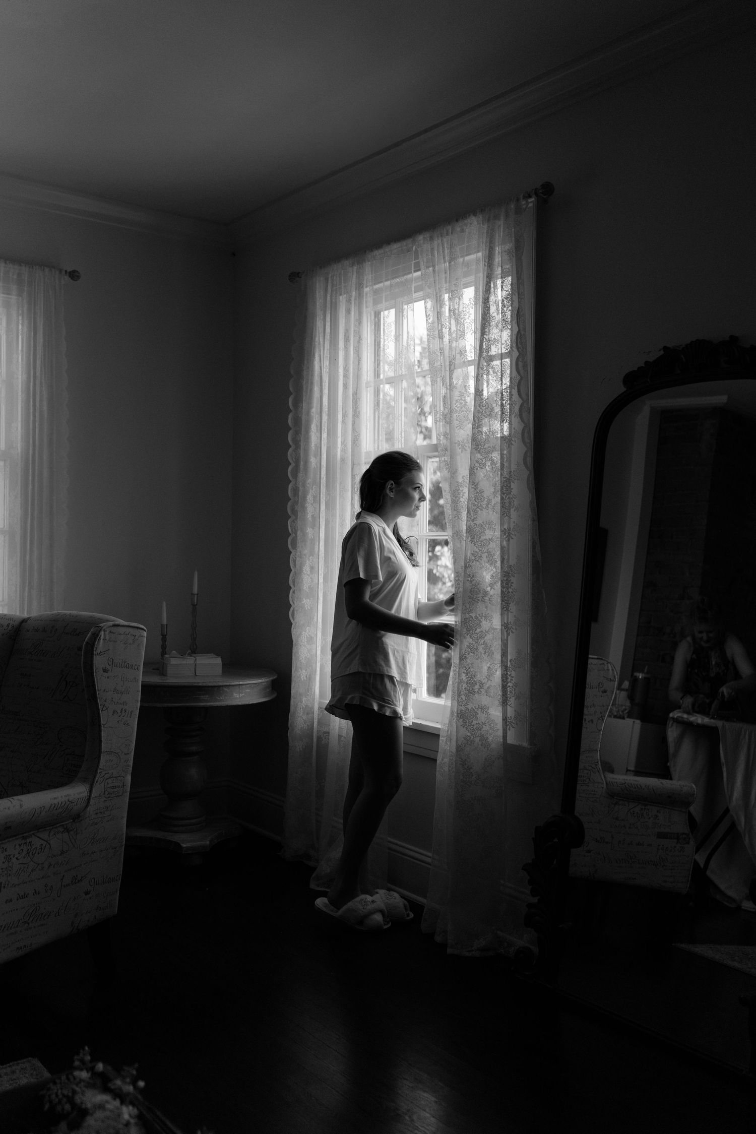 Silhouette by a window in a moody black and white photograph with lace curtains.