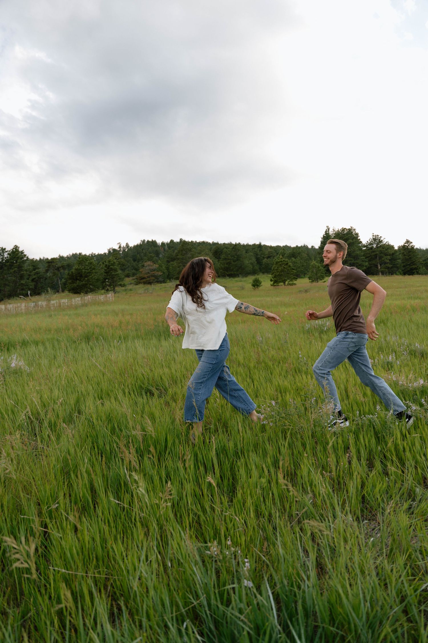 A playful couple runs through a green mountain meadow on a cloudy summer day.
