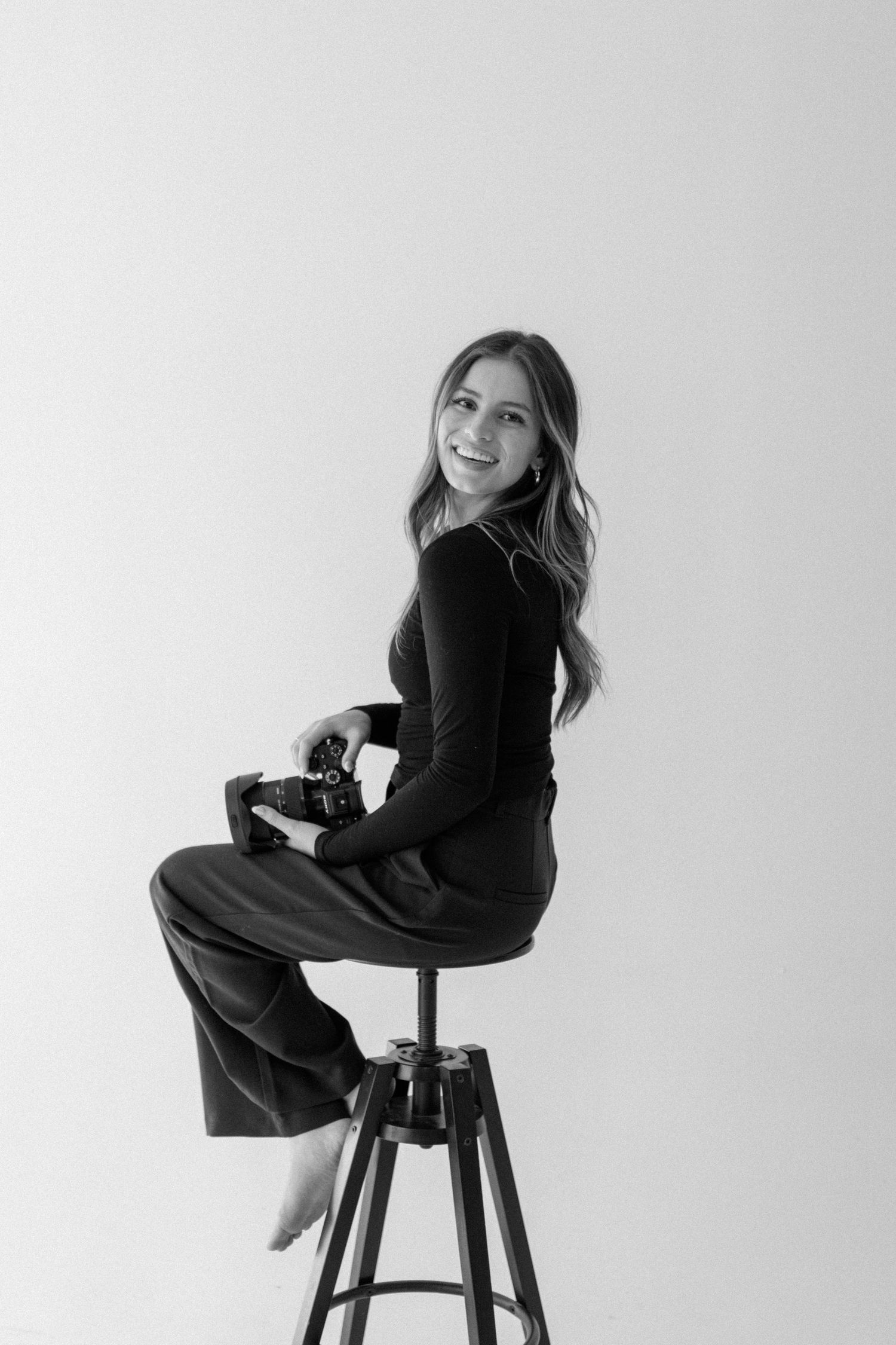 Black and white portrait of a smiling woman sitting on a stool holding a camera, wearing dark outfit.