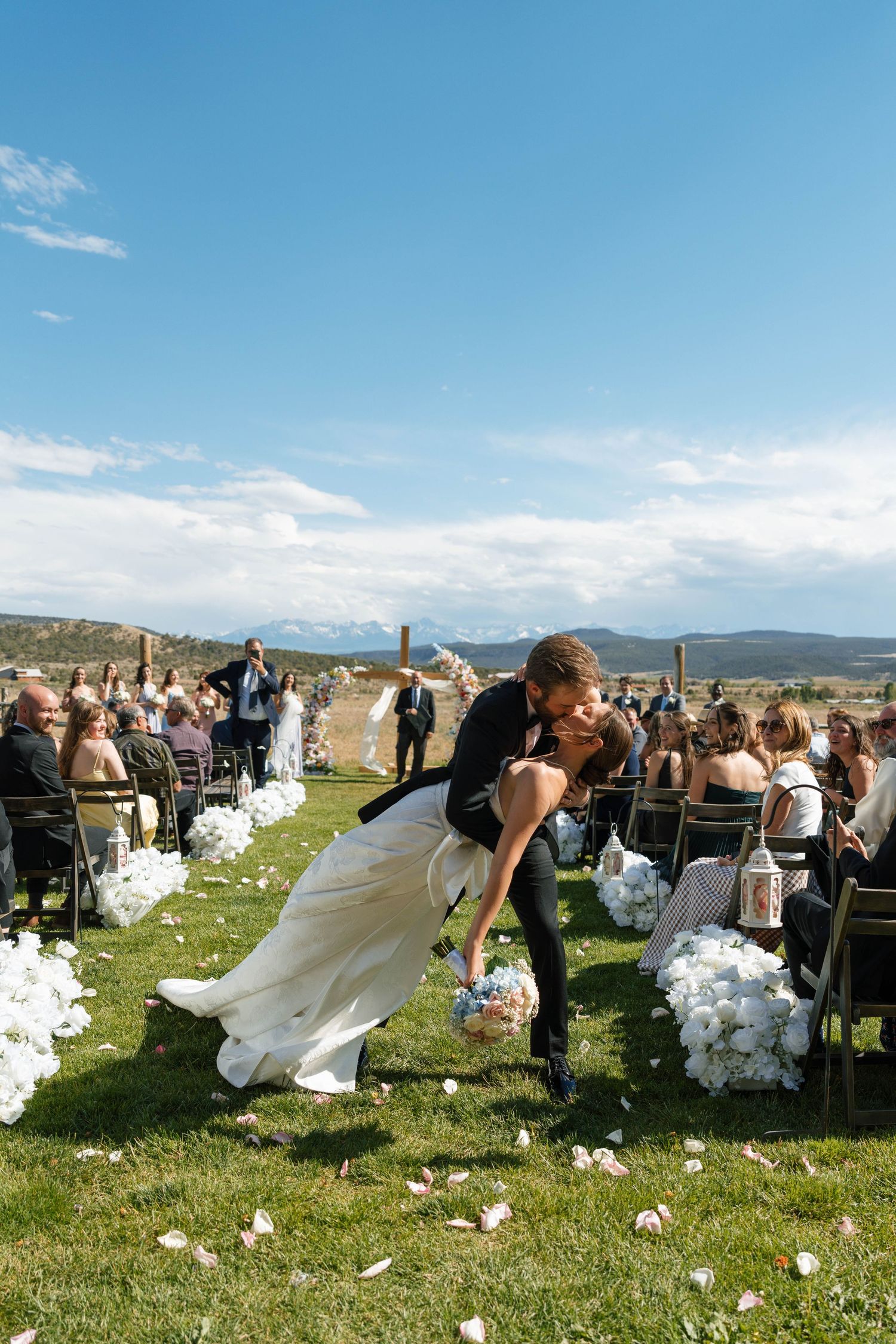 Couple shares romantic dip kiss at outdoor wedding ceremony aisle with mountain views and white floral decor.