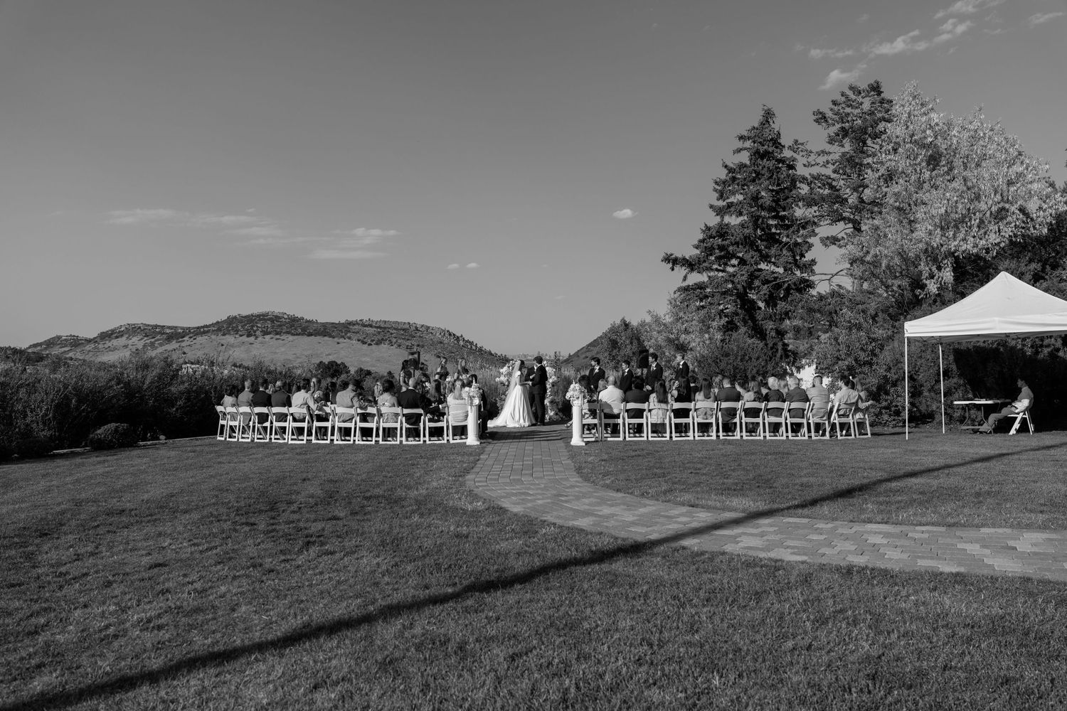 Black and white outdoor wedding ceremony on a lawn with mountain backdrop, guests seated in rows under clear sky.